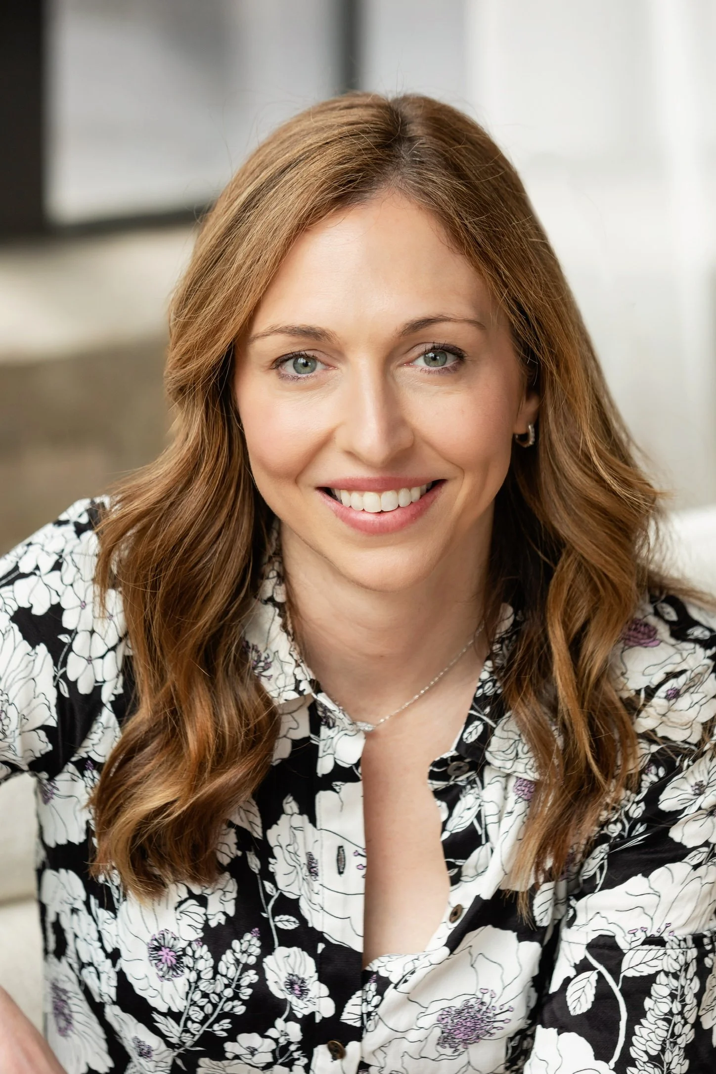 A woman with long, wavy, light brown hair, wearing a black and white floral-patterned shirt, smiling with visible teeth, in an indoor setting with a blurred background.