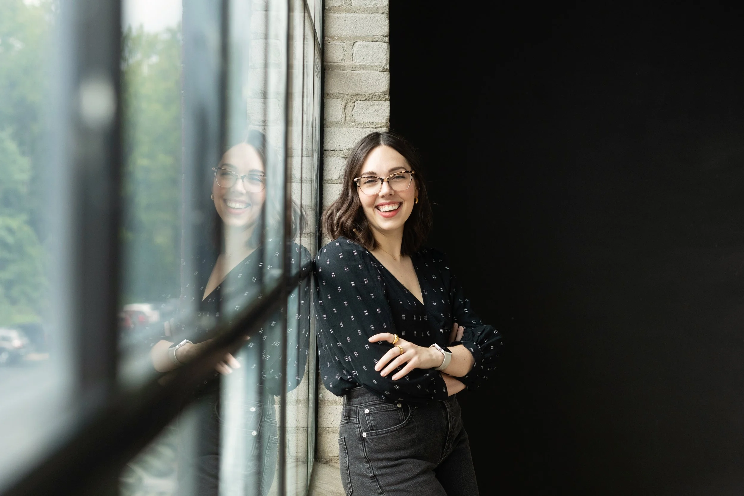 A young woman with glasses and shoulder-length dark hair smiling, standing by a large window with her arms crossed, with her reflection visible on the glass. She is wearing a black patterned blouse and dark jeans, in an industrial-style setting with 