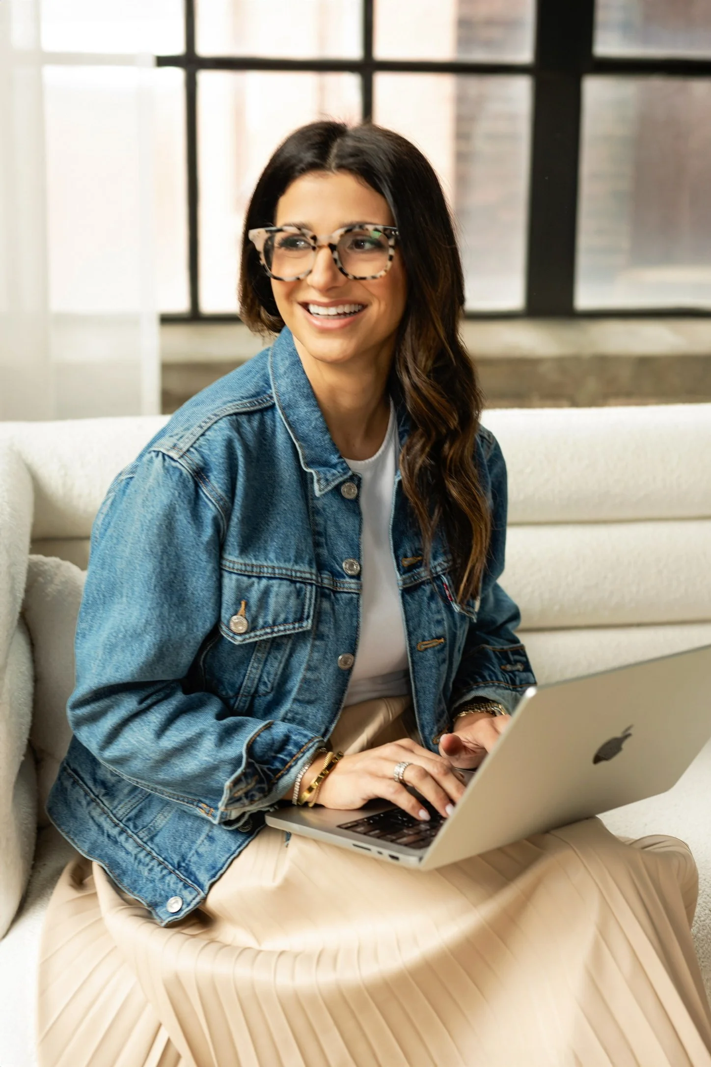 A woman with glasses and dark hair sitting on a sofa with a laptop, smiling and looking to the side, in a bright room with large windows.