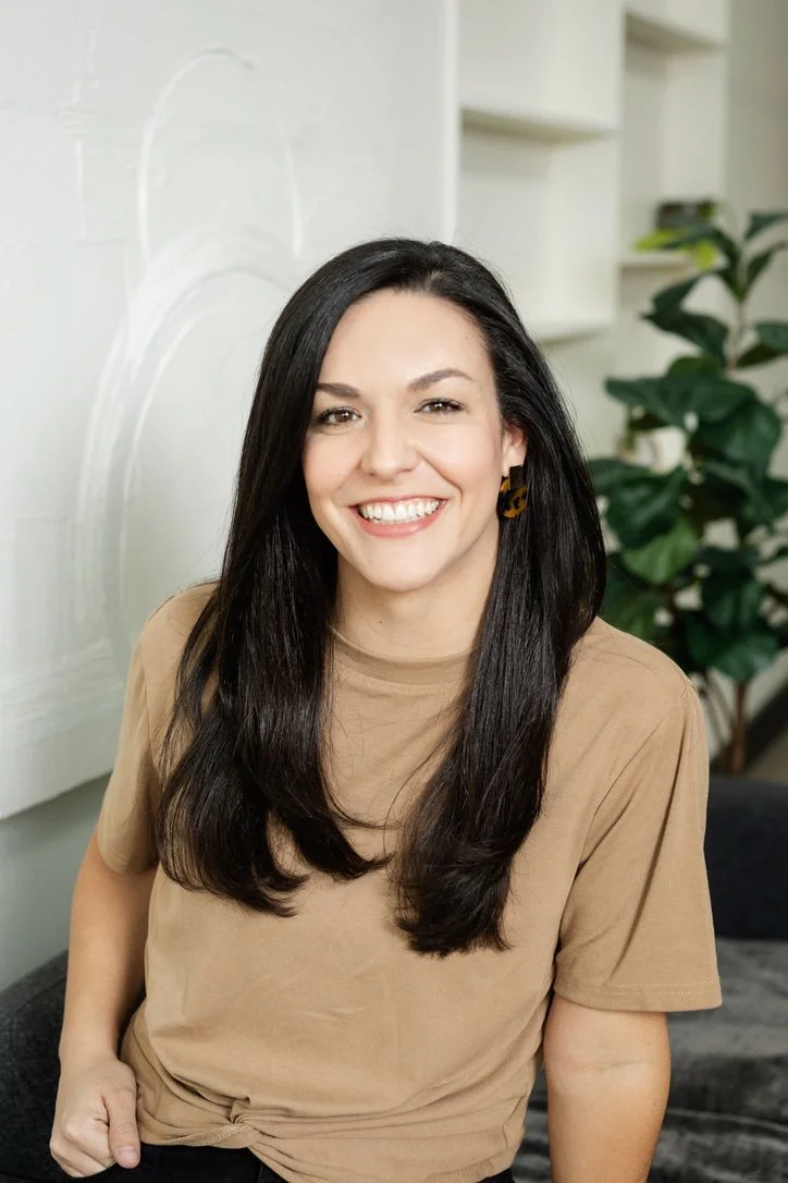 A smiling woman with long dark hair, wearing a beige t-shirt, sitting indoors with a green plant and shelves in the background.