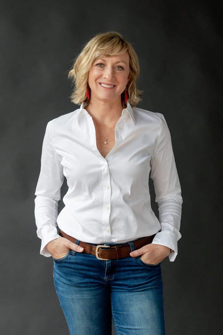A smiling woman with blonde hair wearing a white blouse, blue jeans, and red earrings, posing with hands in pockets against a dark background.
