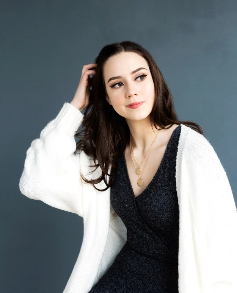 A young woman with long dark hair, wearing a dark dress and a white cardigan, poses against a plain background.