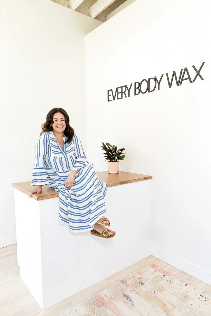 Woman wearing a blue and white striped dress sitting on a wooden counter with a potted plant, smiling at the camera in front of a white wall with the sign 'Every Body Wax'.