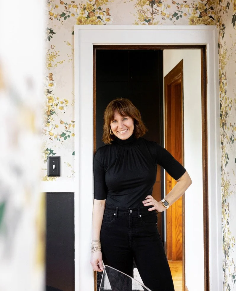 A woman with shoulder-length brown hair, wearing a black turtleneck and black jeans, smiling and standing in a doorway inside a home with floral wallpaper.