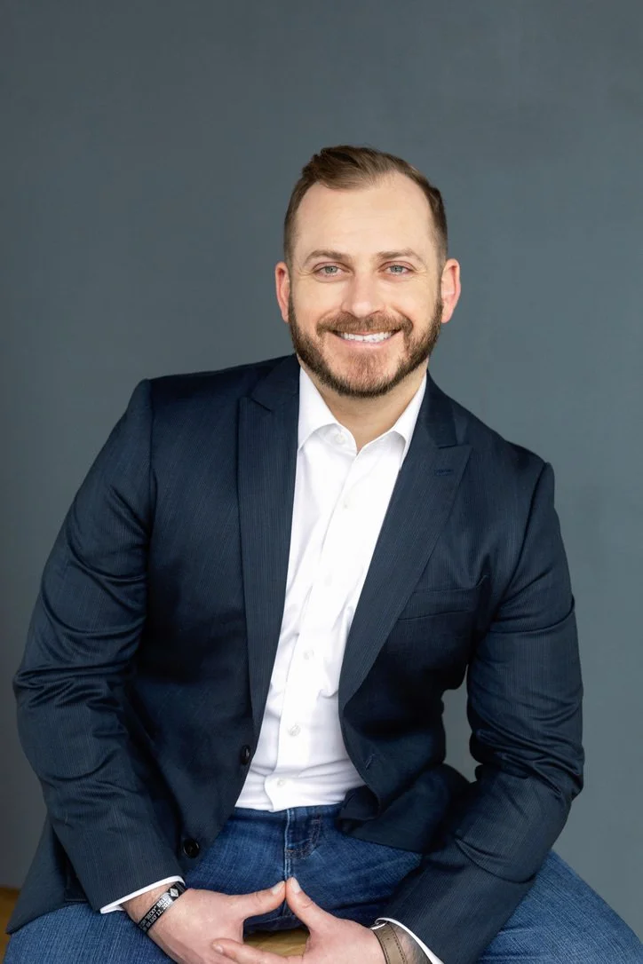 Portrait of a smiling man wearing a navy blazer, white shirt, and blue jeans against a dark gray background.