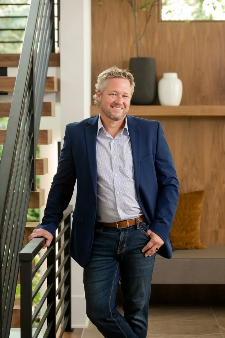 A man with curly gray hair and a beard, wearing a blue blazer, light-colored shirt, and jeans, smiling and leaning against a staircase railing in a modern interior.