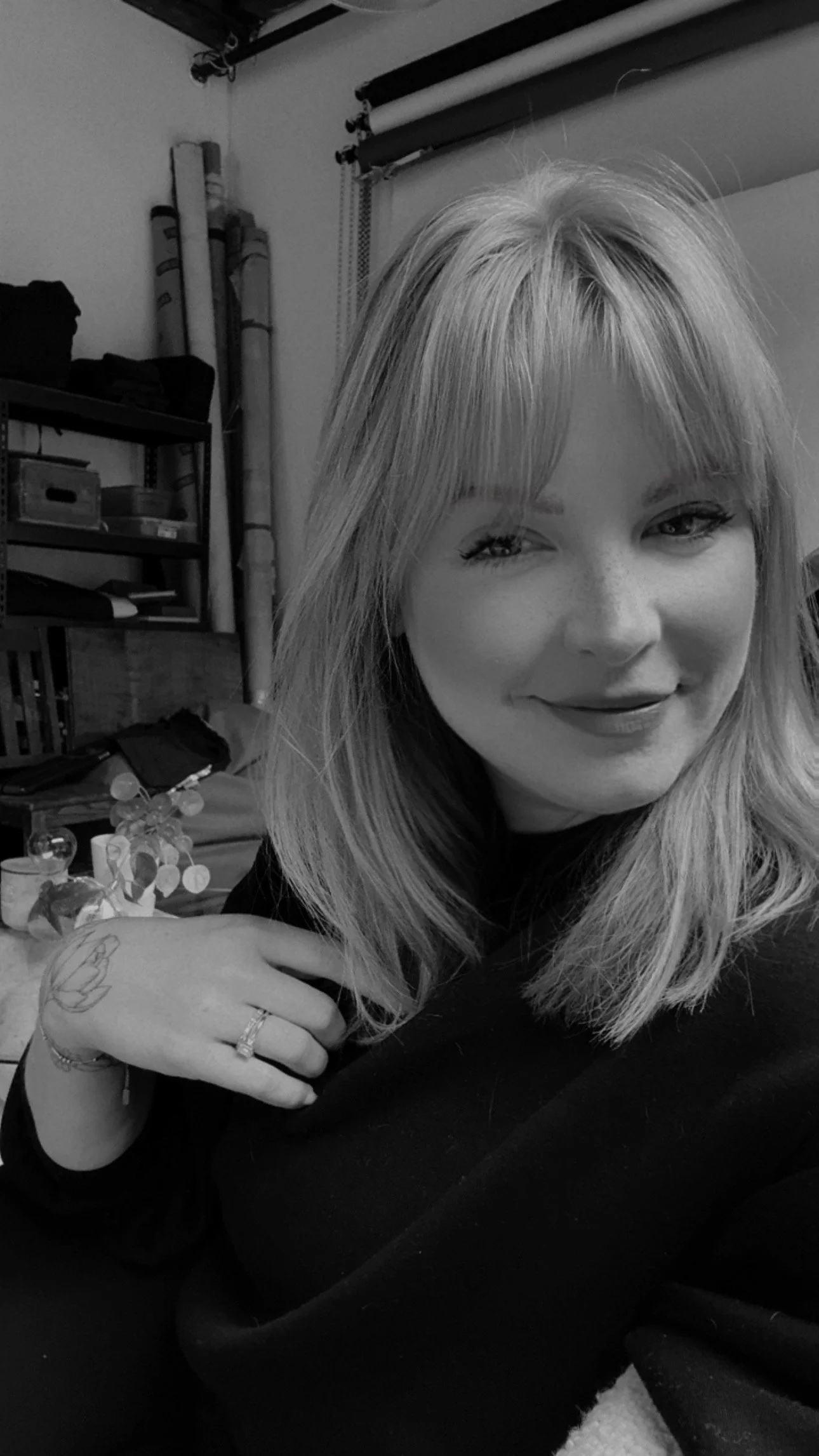 A woman with blonde hair and a tattoo on her left hand, smiling softly at the camera, in a room with photography equipment, shelves, and rolled backdrop papers in the background.