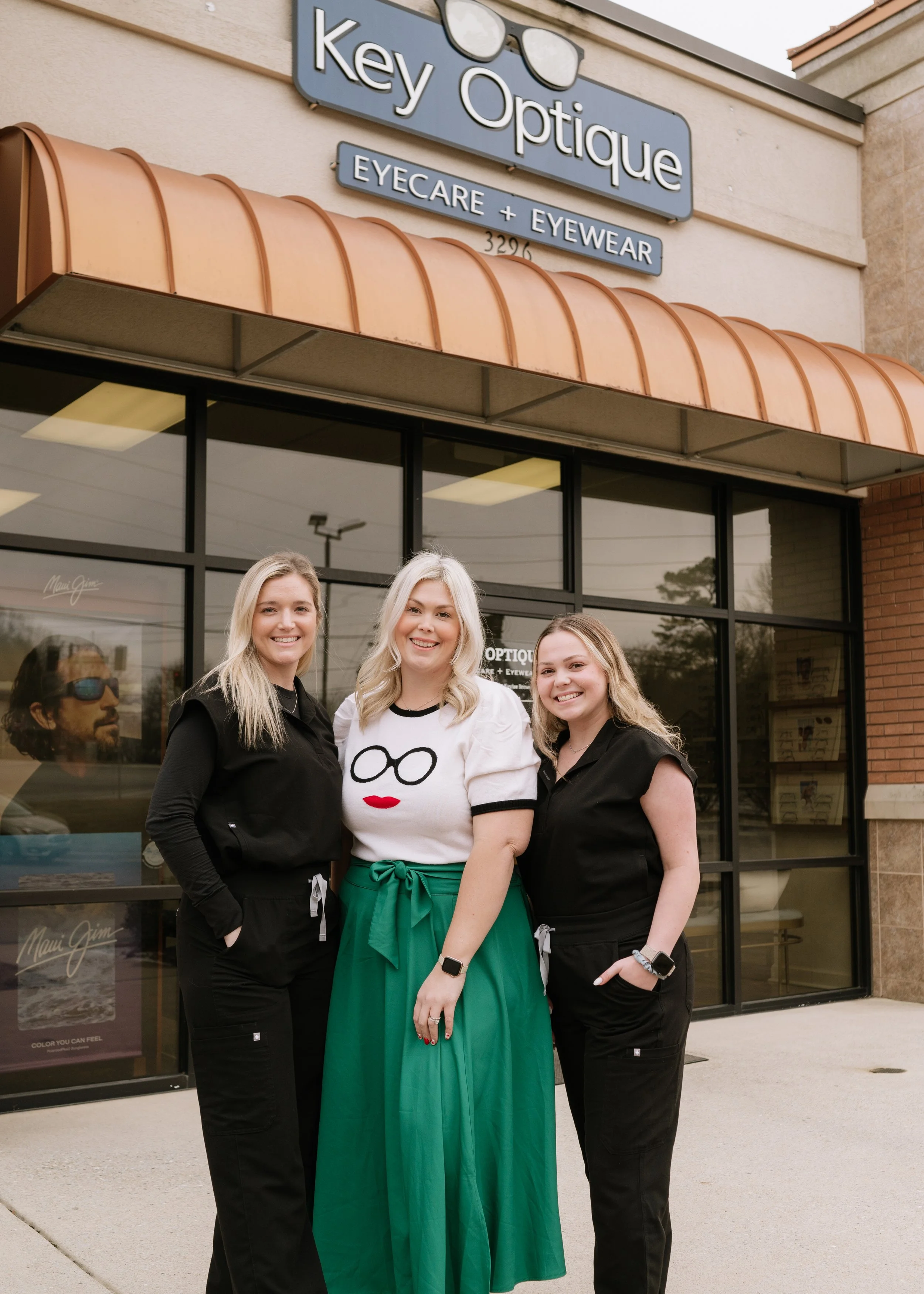 Three women standing in front of Key Optique eyeglass store, smiling for the camera.