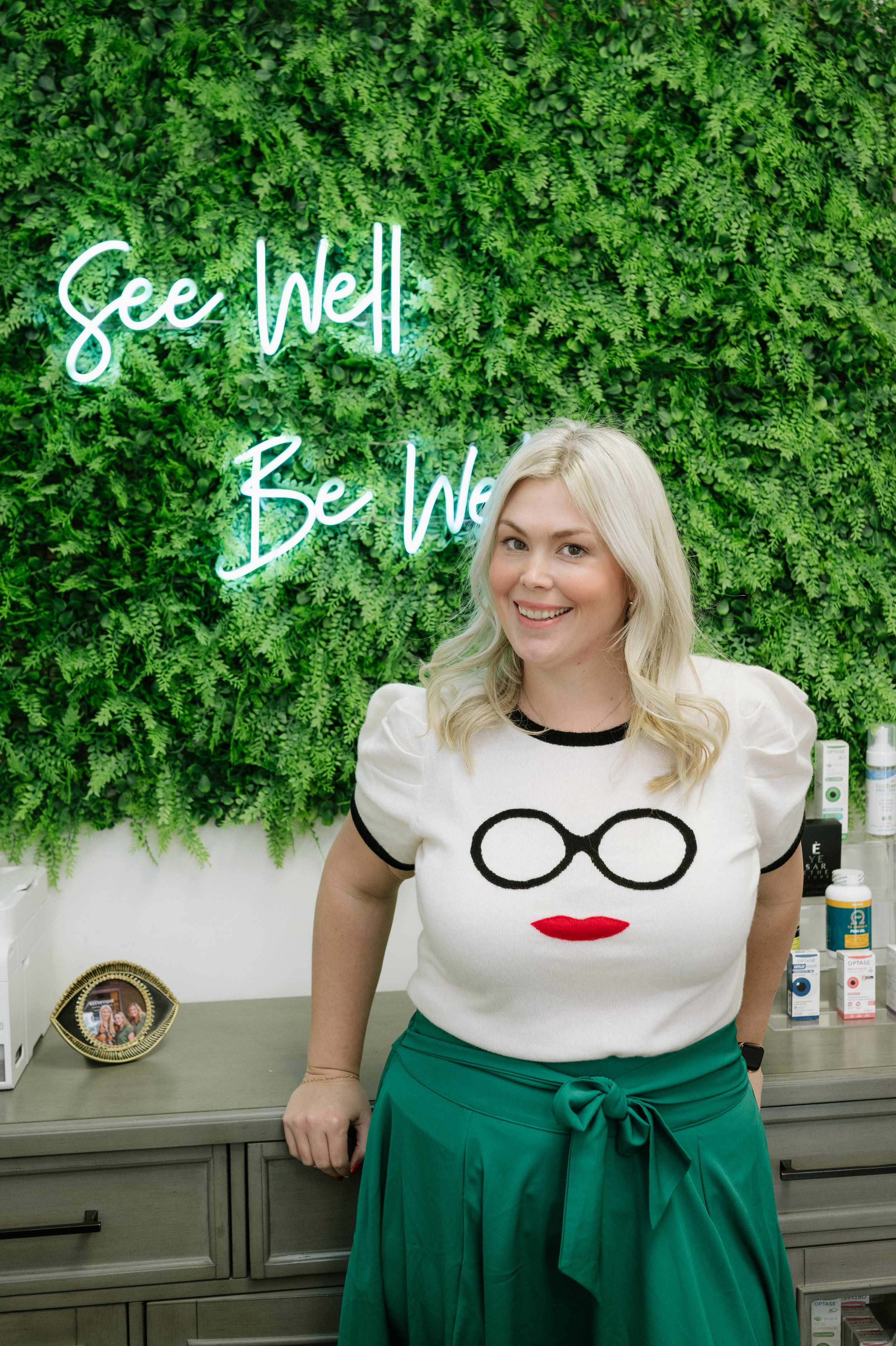 A woman with blonde hair, wearing a white sweater with glasses and lips printed on it, standing in front of a green wall with a neon sign that says 'See Well, Be Well'.