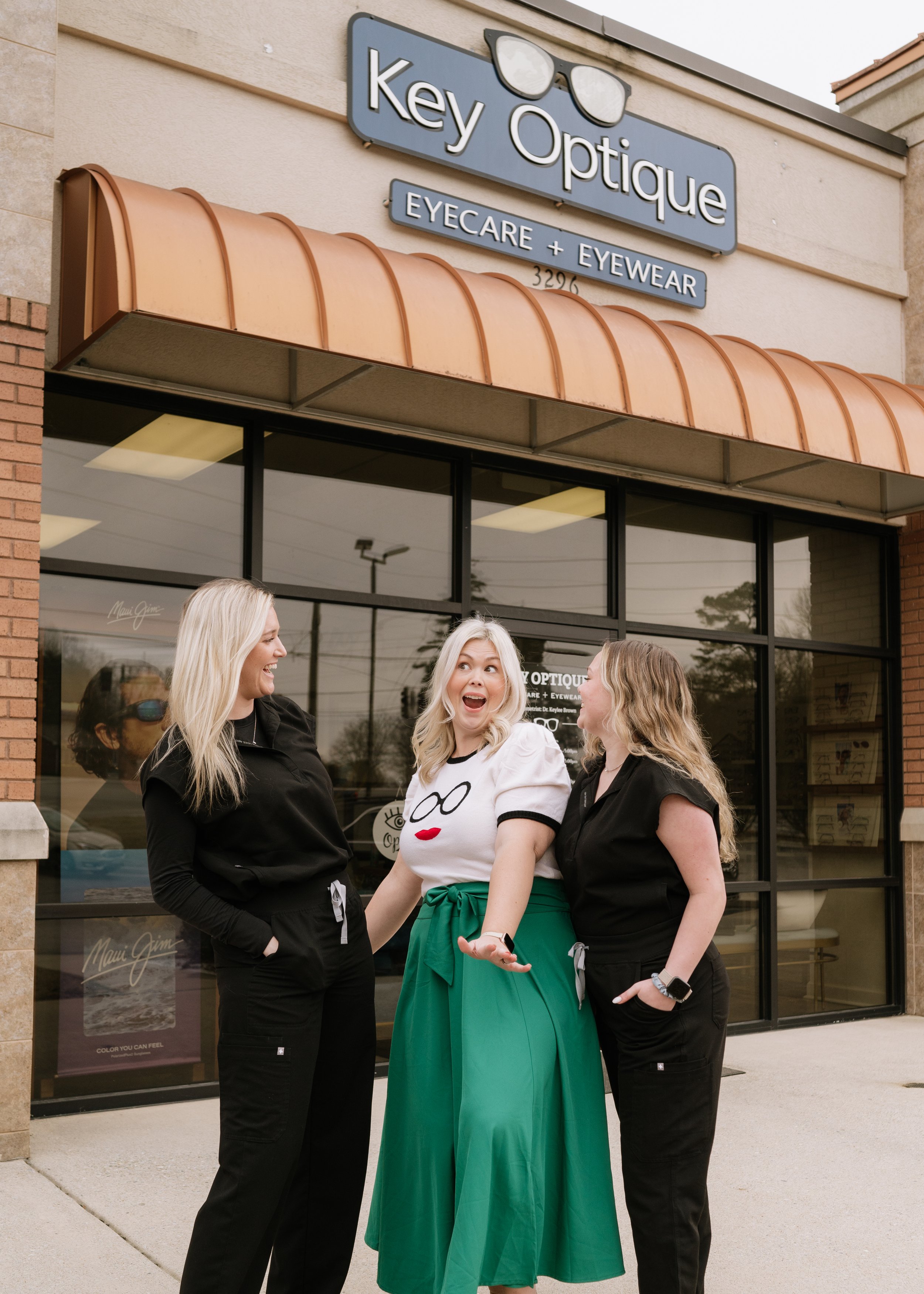 Three women standing outside Key Optique eyeglasses store, smiling and talking, with the store's sign visible above them.
