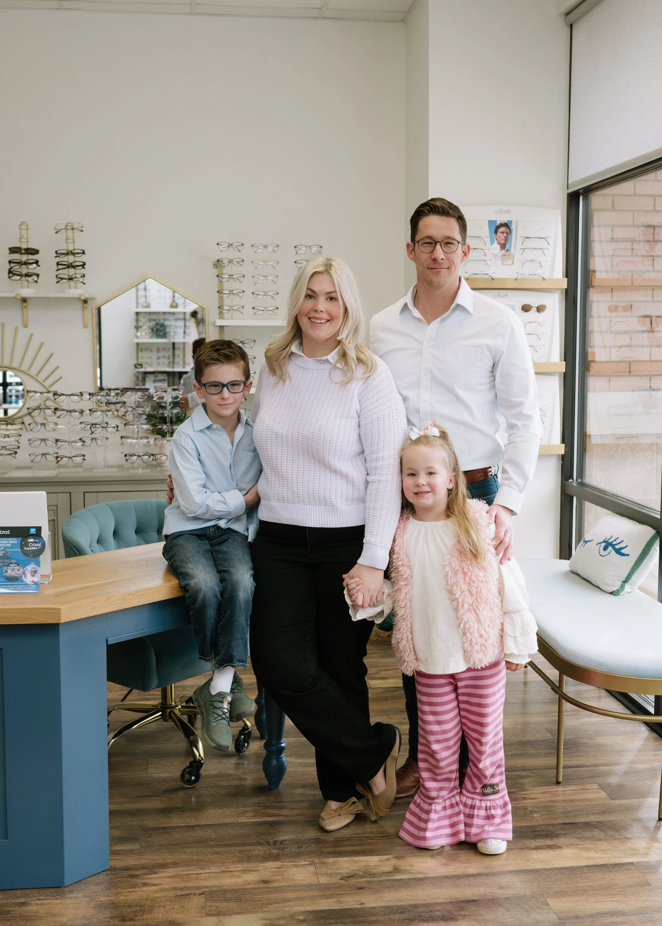 A family of five in an eyewear store, with eyeglasses displays on the wall behind them. The children are sitting and standing near the counter, and the parents are in the center.