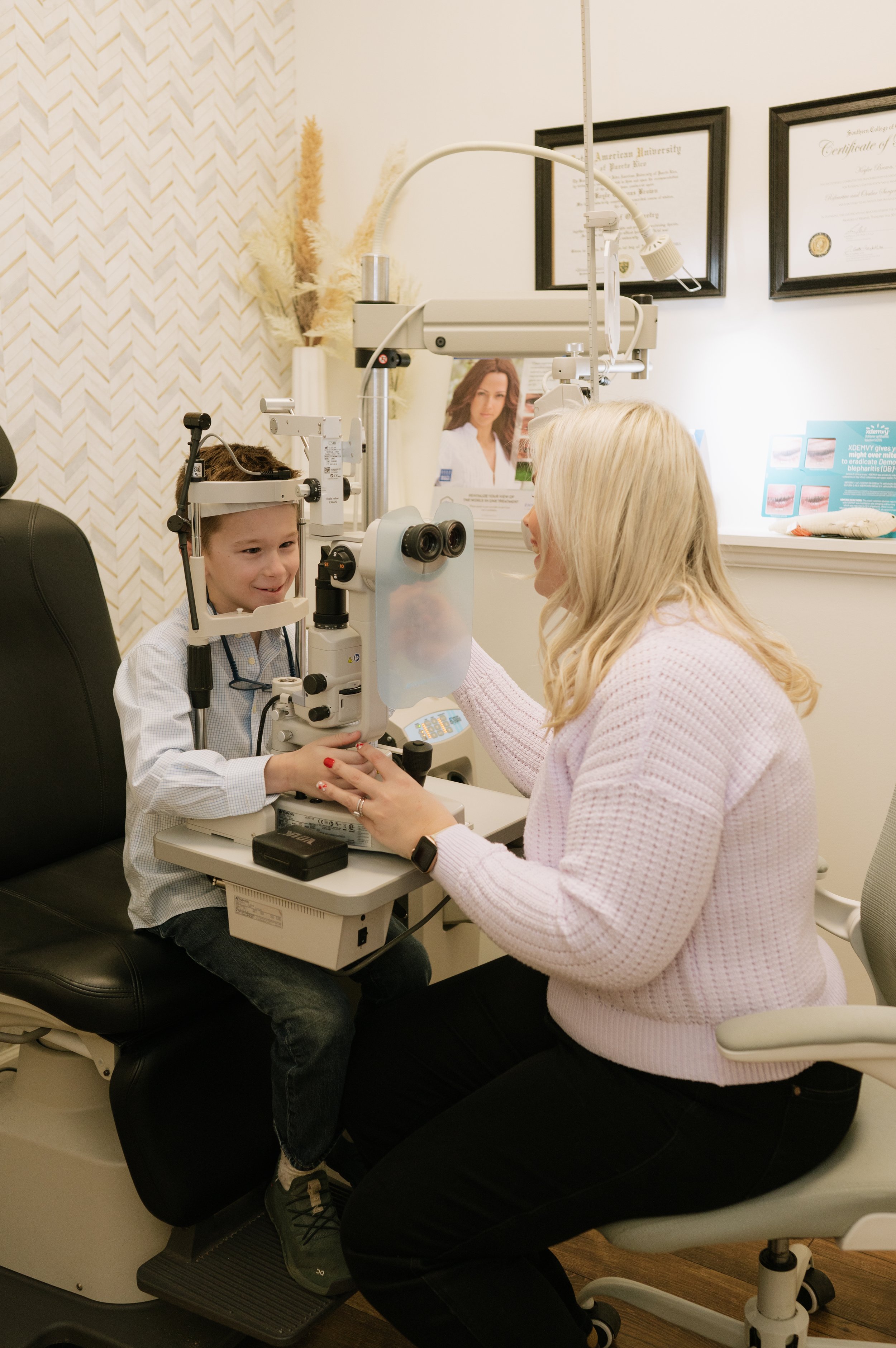 A young boy visits an eye doctor, sitting at a slit lamp exam table with a female optometrist, as she checks his vision.