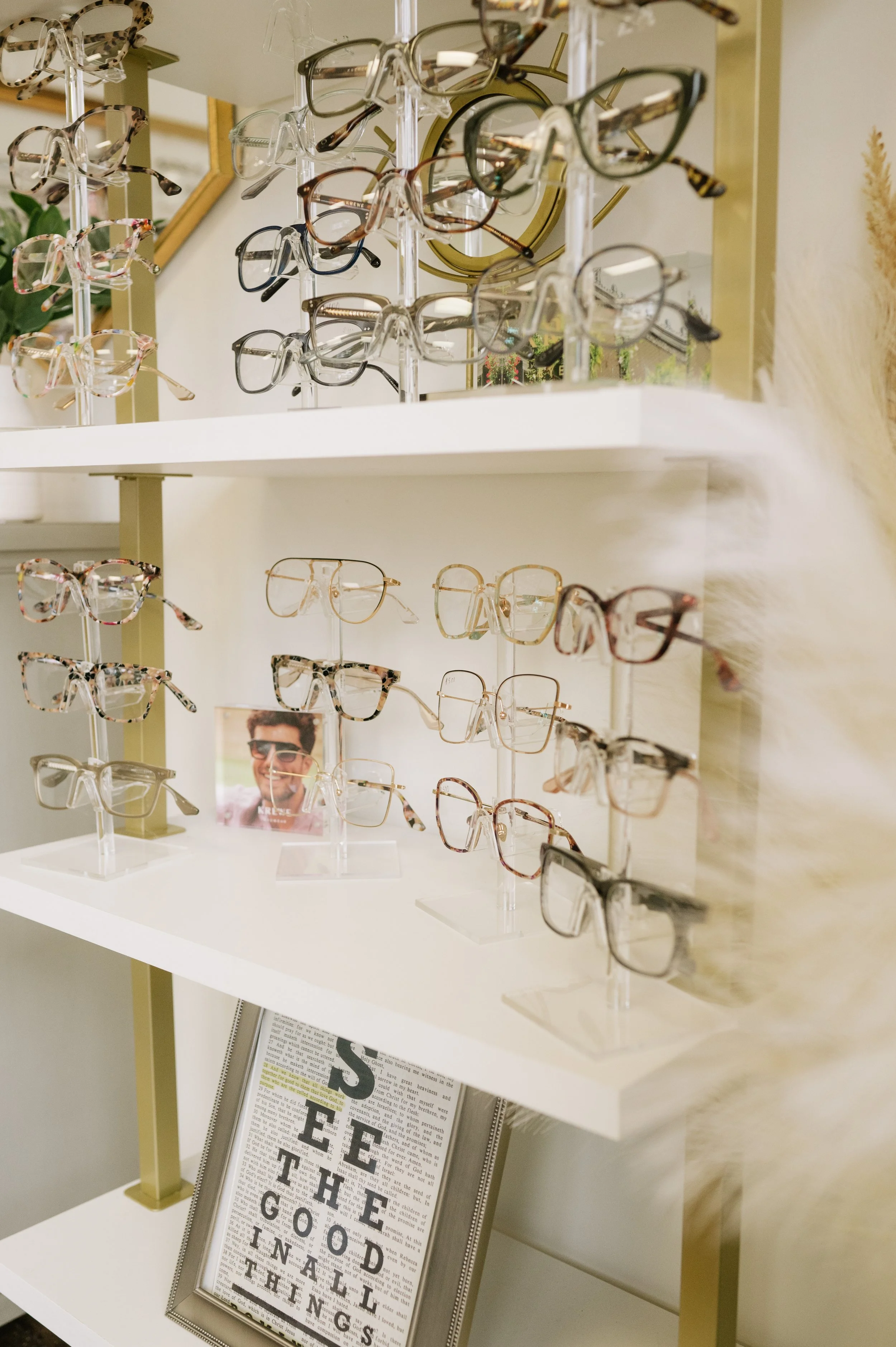 Display of various eyeglasses on clear stands in an optometry shop, with a framed quote on the lower shelf that reads "See the good in all things."