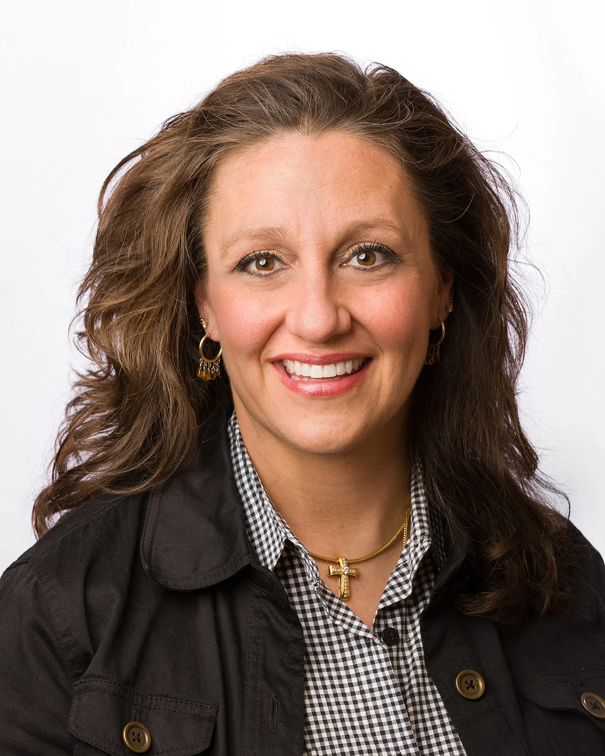 A woman smiling with brown, wavy hair, wearing a black jacket over a checkered shirt, and a gold cross necklace. She also has earrings and a small hoop earring in each ear.