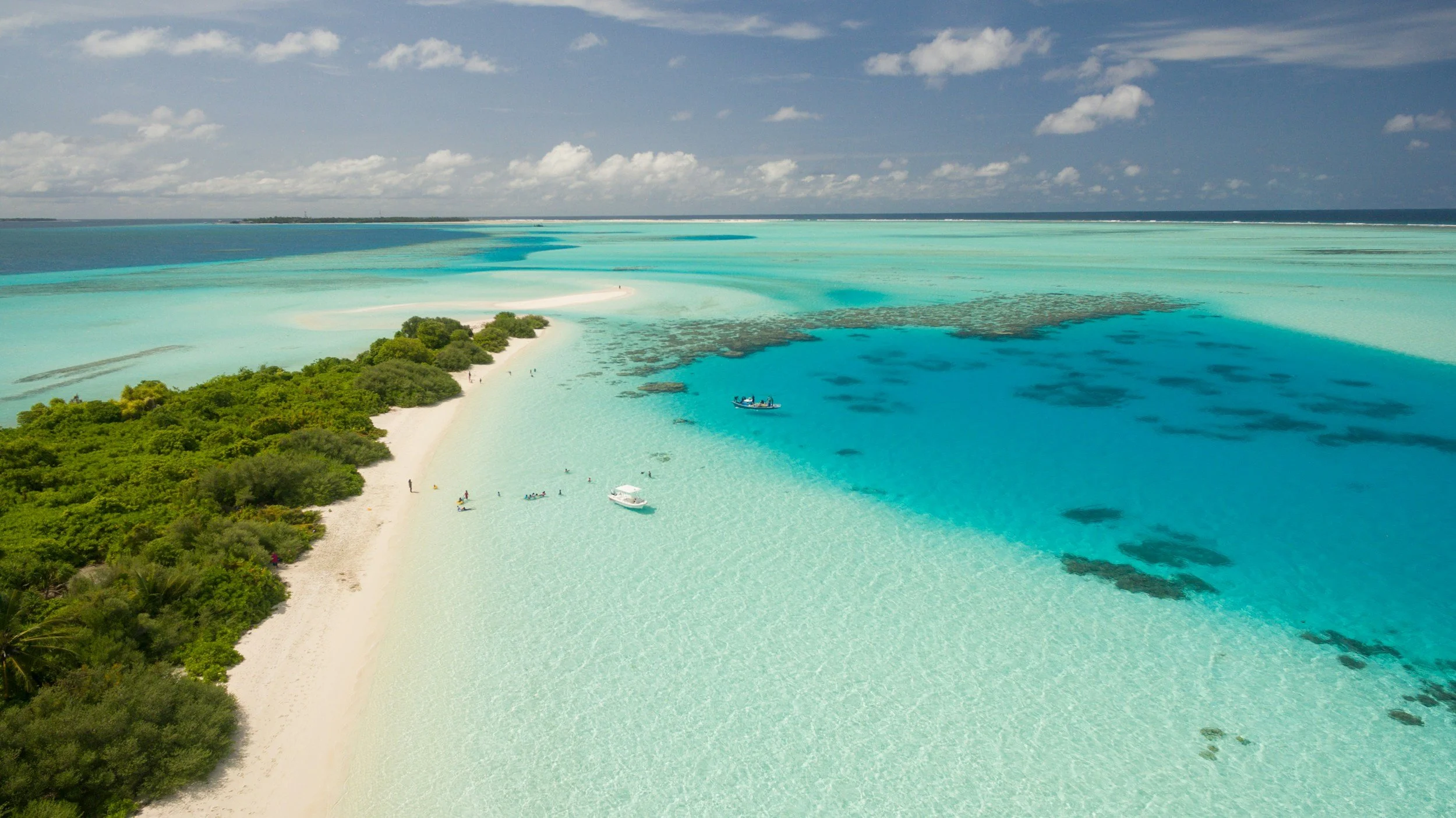 Aerial view of a tropical beach with white sand, lush green trees, and turquoise water with dark patches of seaweed or coral, under a partly cloudy sky with small white clouds.