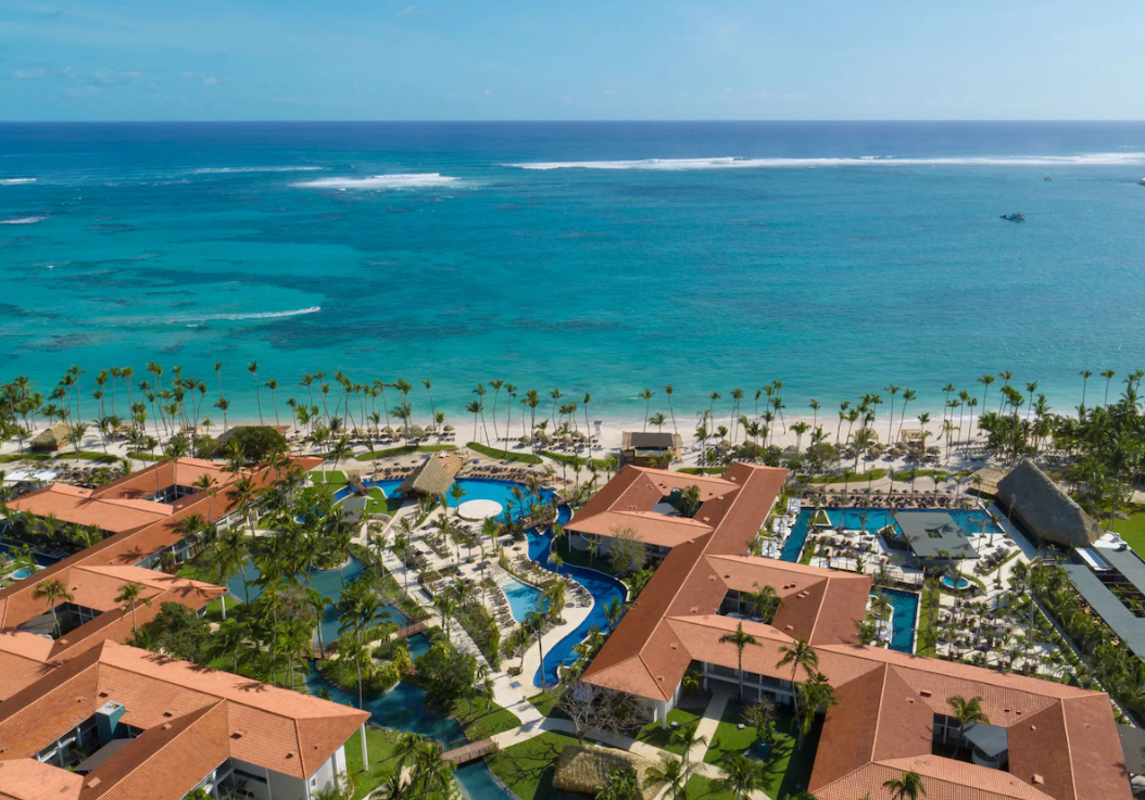 Aerial view of a tropical beach resort with swimming pools, palm trees, and beach access to the ocean.