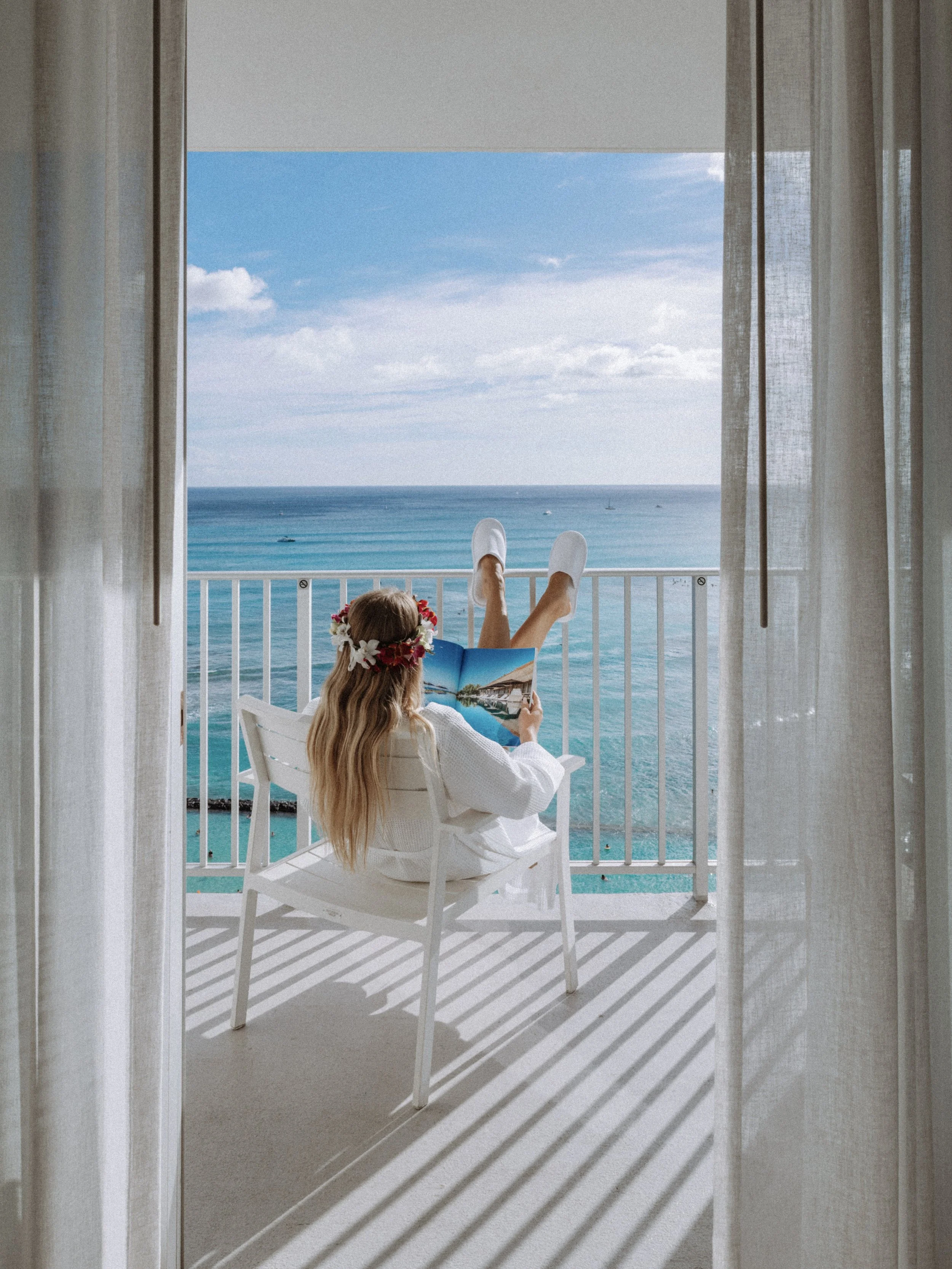 A woman with long blonde hair wearing a white robe and a flower crown is sitting on a white chair on a balcony, reading a magazine. She has her feet up on the balcony railing, overlooking the ocean. The scene is framed by white sheer curtains and sunny weather.