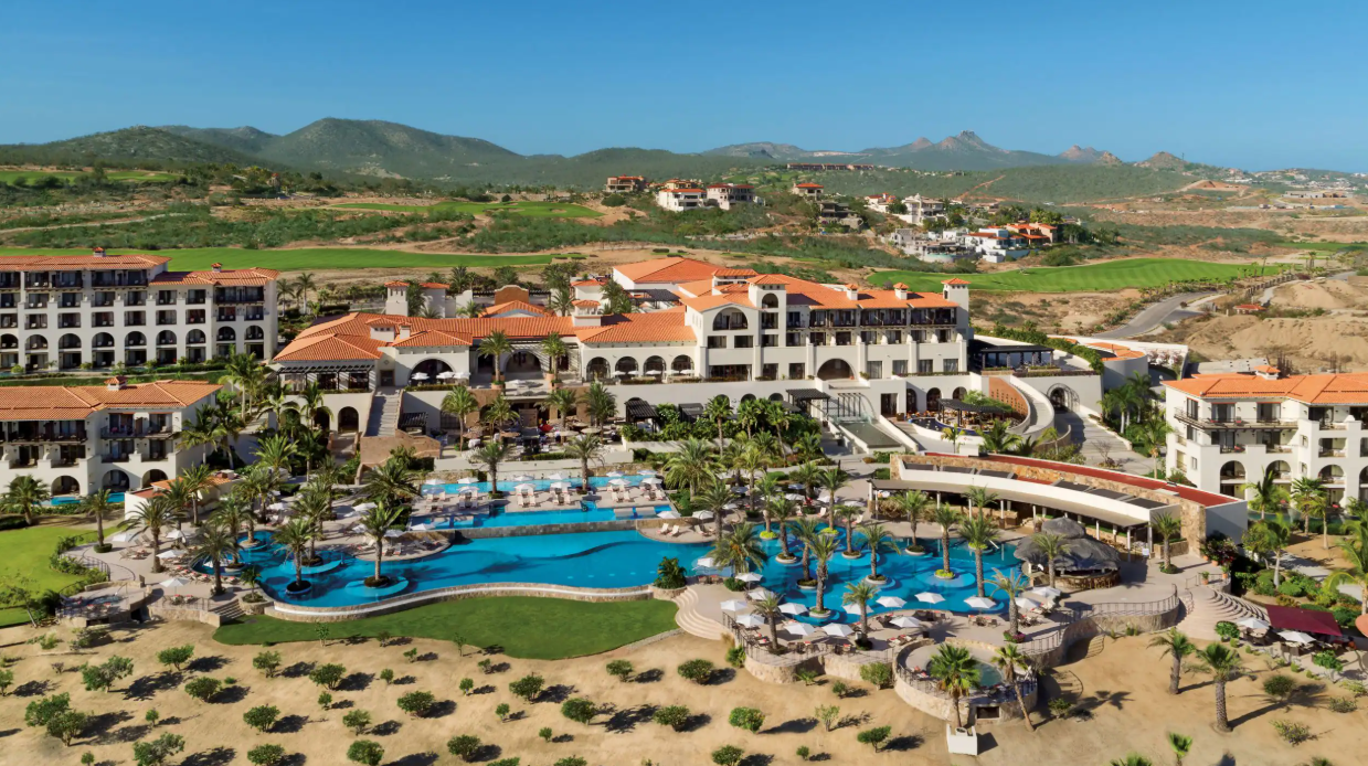 A luxurious resort with multiple buildings featuring terracotta roofs, surrounded by palm trees and lush landscaping, with a large swimming pool area in the foreground, set in a mountainous landscape under a clear blue sky.