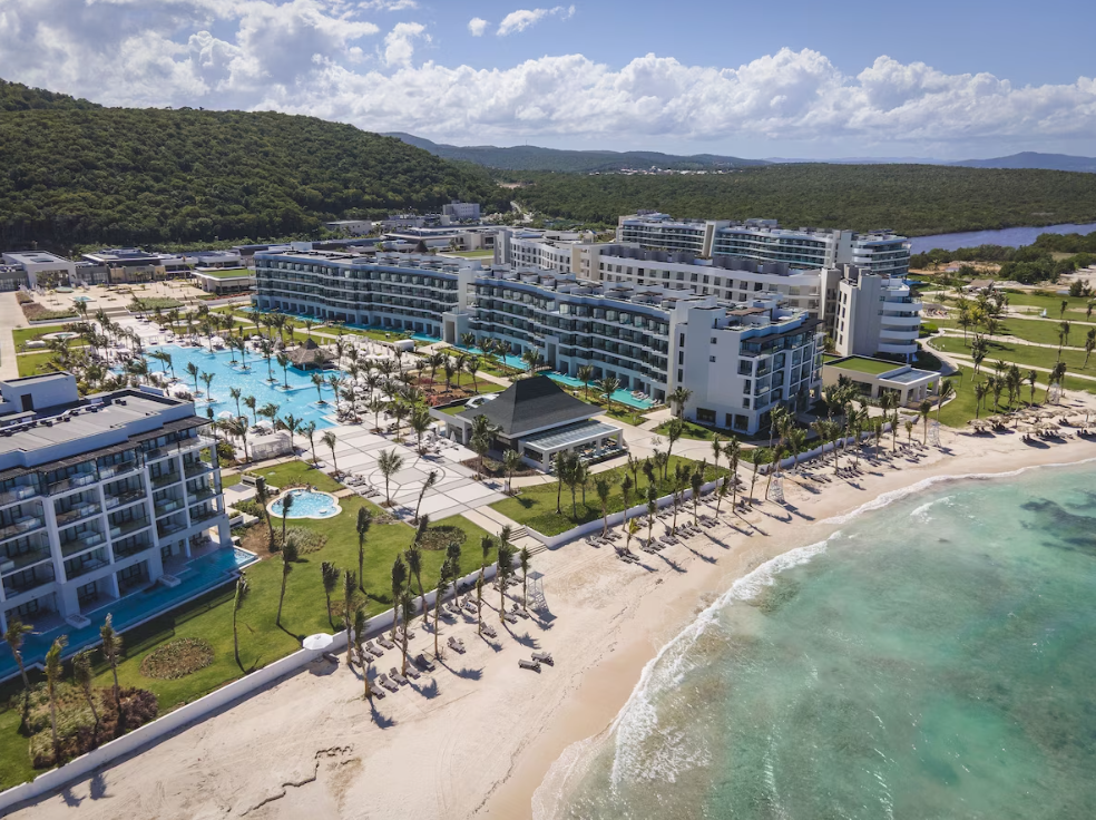 Aerial view of a beachfront resort with multiple modern white buildings, a large swimming pool, and palm trees, alongside a sandy beach and ocean, with green hills and a body of water in the background.