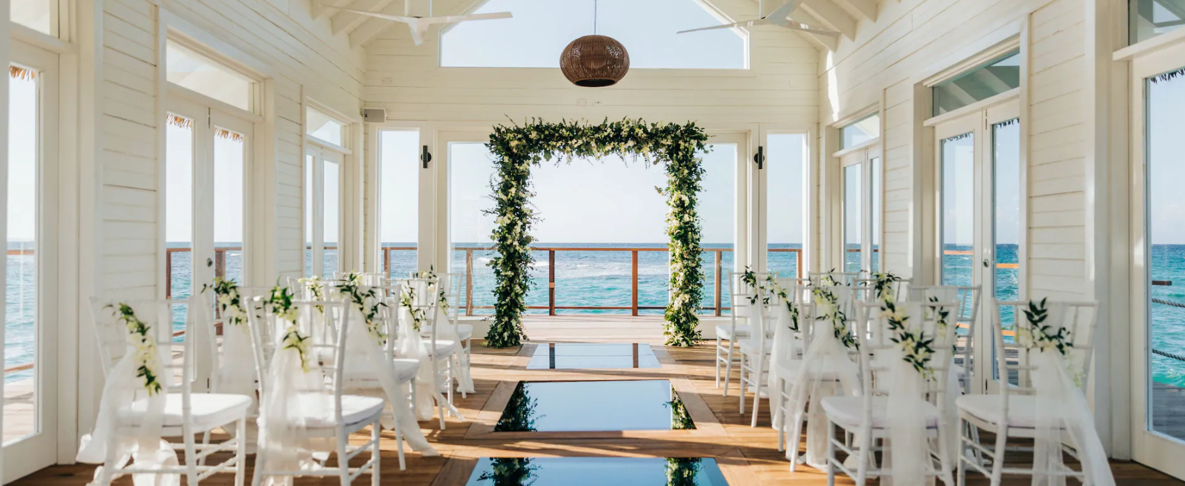 Beachside wedding setup in a white enclosed pavilion with glass walls, decorated with a floral arch, white chairs with green foliage, and overlooking the ocean.