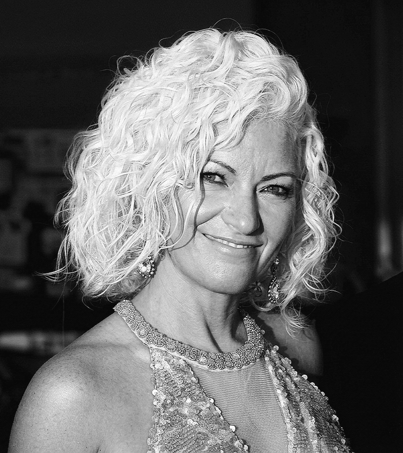 A woman with curly blonde hair, wearing earrings and a sparkly dress, smiling at the camera.