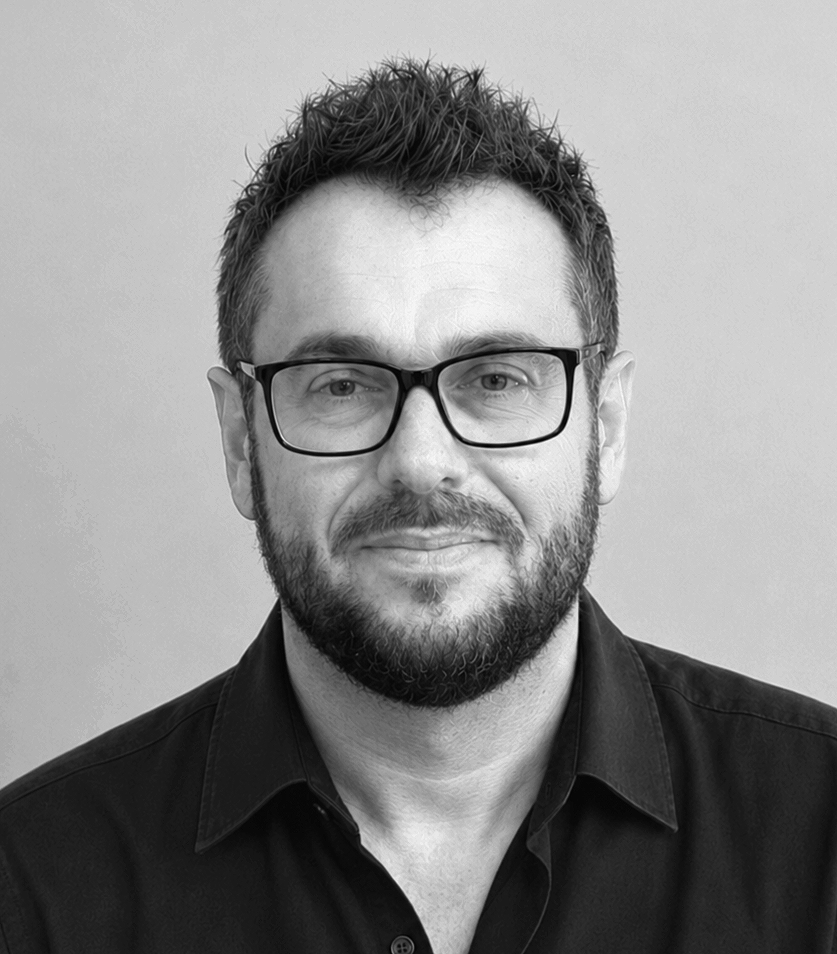 Black and white portrait of a man wearing glasses and a collared shirt, smiling slightly, with short curly hair and a beard.