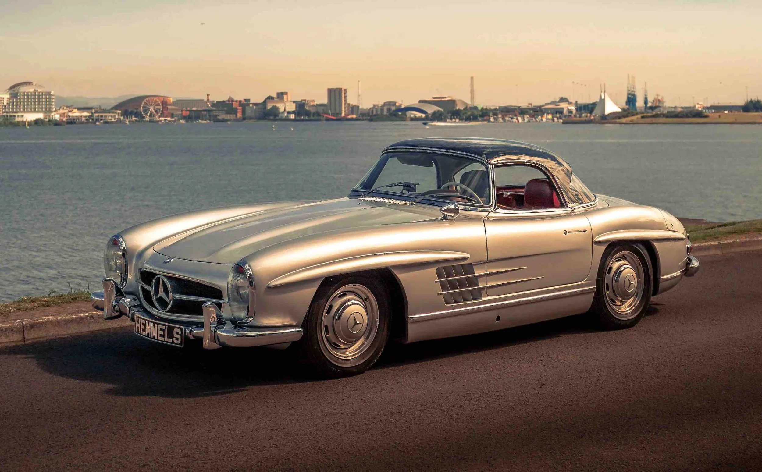 A vintage silver Mercedes-Benz car parked near a body of water with city buildings, a ferris wheel, and cranes in the background.