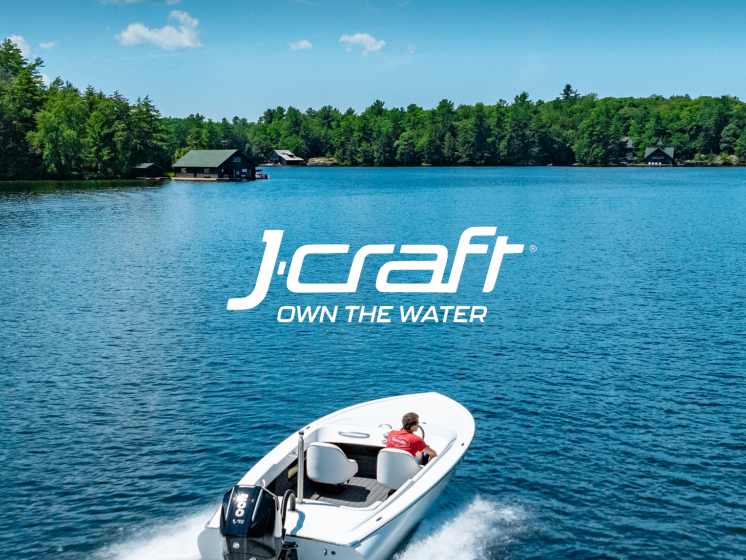 A person in a red shirt rides a white boat on a calm blue lake surrounded by green trees and houses, with the J.Craft logo and the slogan 'Own the Water' in the center.