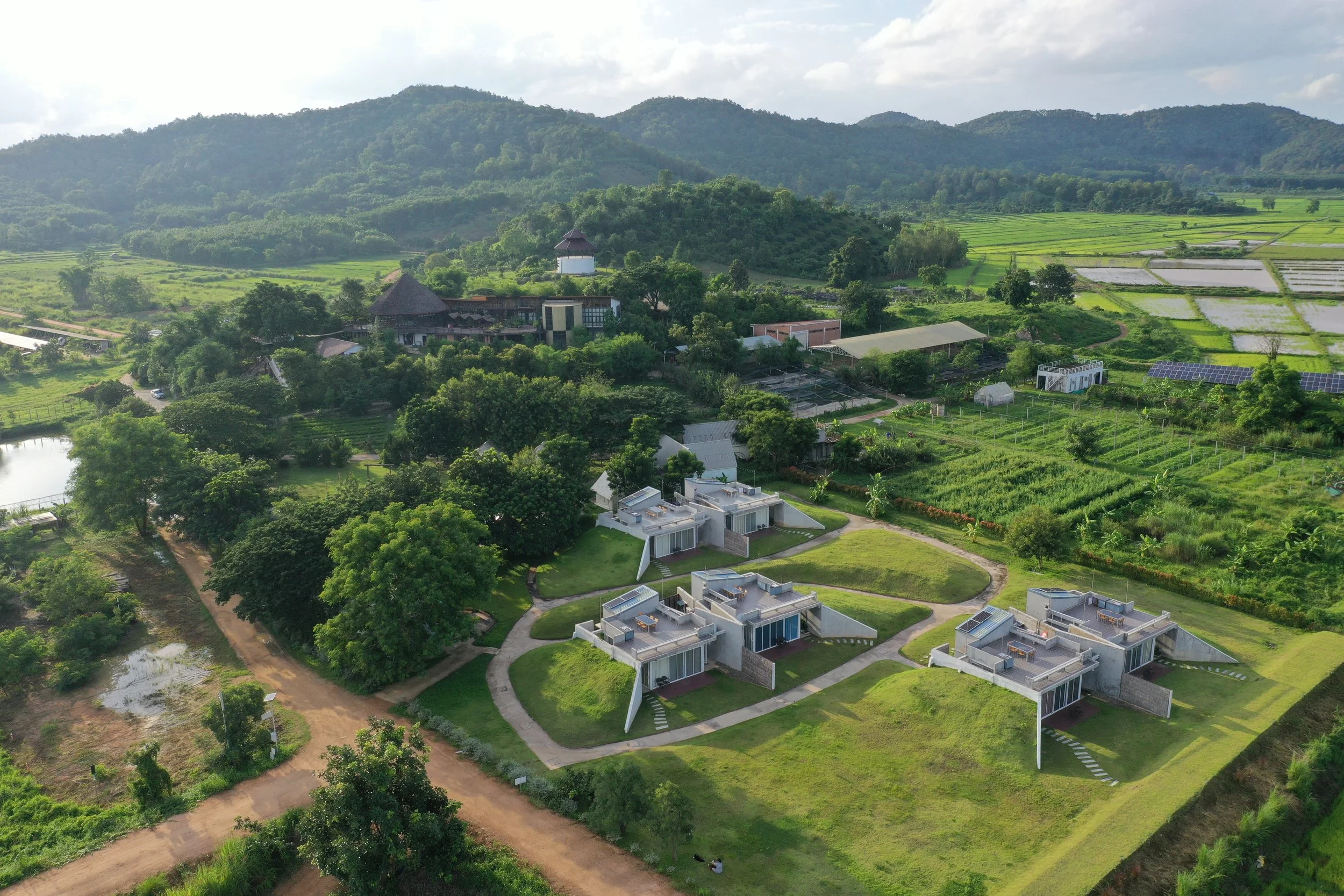 Aerial view of modern houses with unique architecture surrounded by lush green landscape, trees, and mountains in the background.