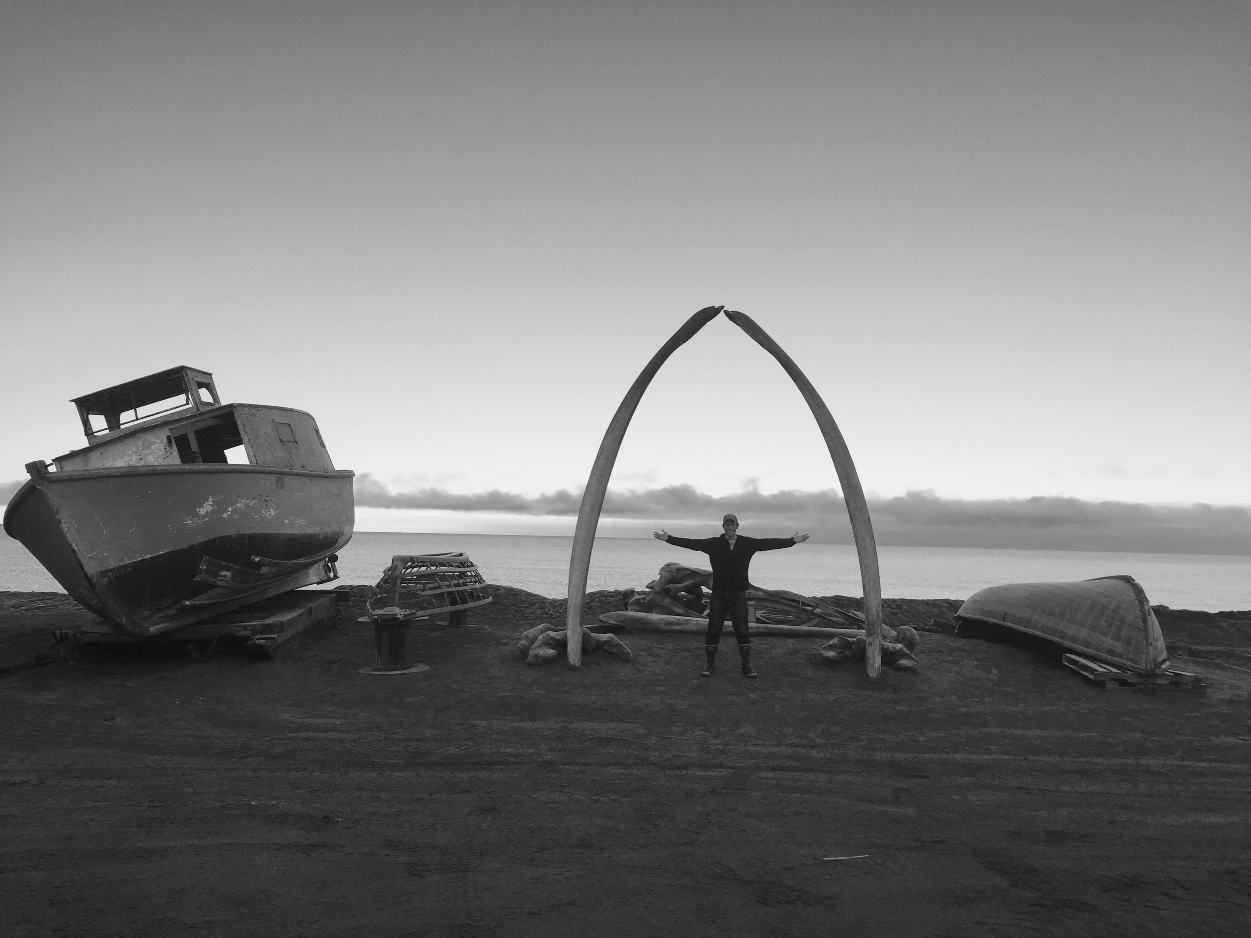 Christian stands beneath the whale bone arch, near several retired wooden boats on the beach of Barrow, Alaska.