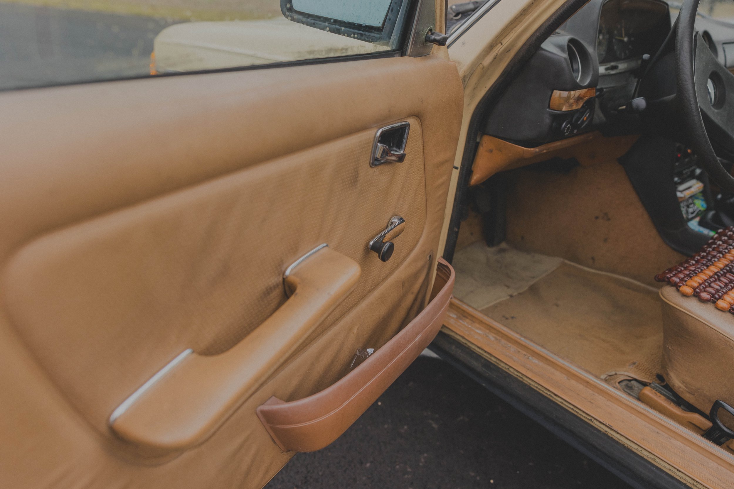 Interior of an old car with open door, beige upholstery, manual window crank, and beaded seat cover.