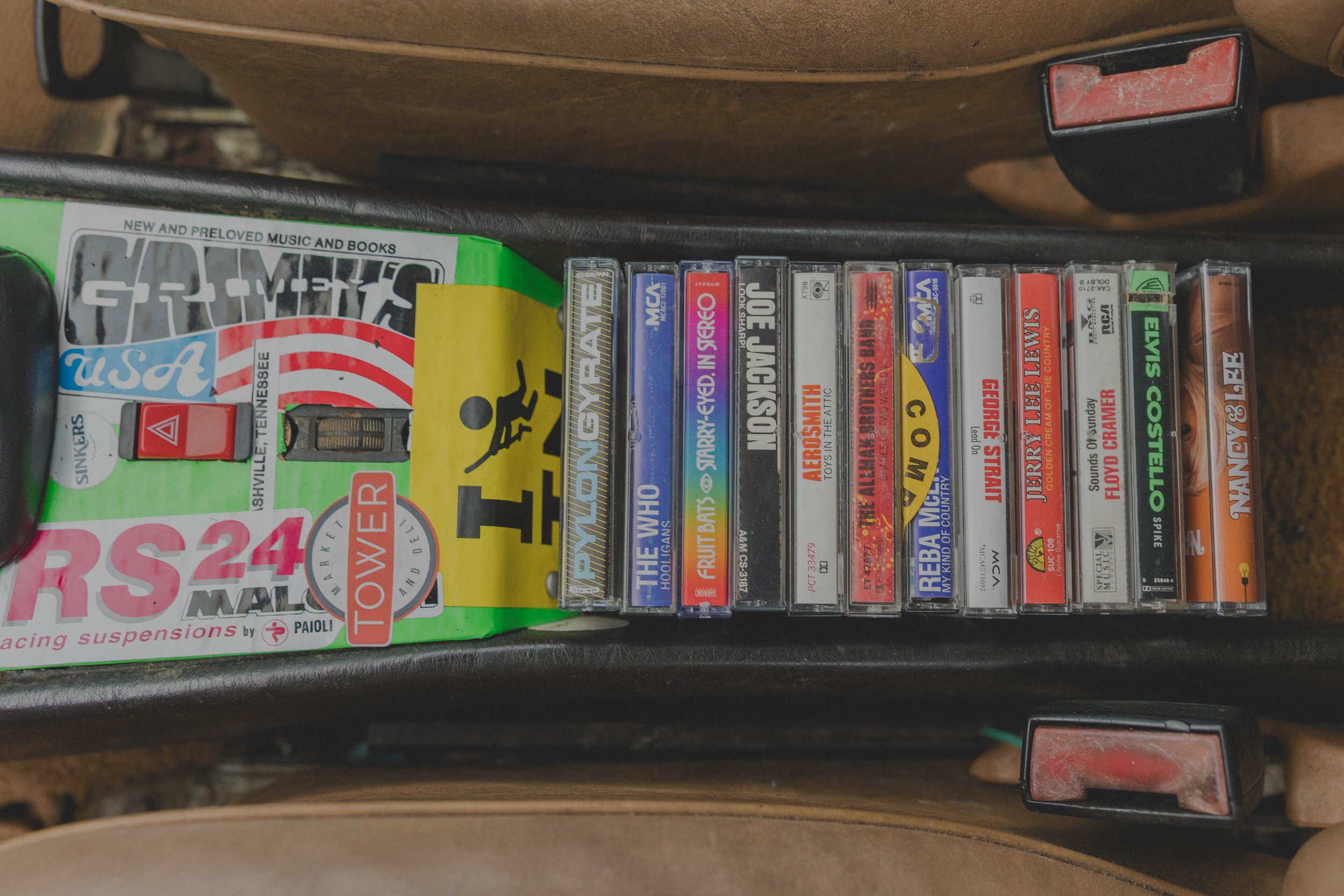 View of a car seat's cassette holder filled with various music tapes, including artists like The Who, Aerosmith, and Elvis Costello, alongside stickers and labels."}