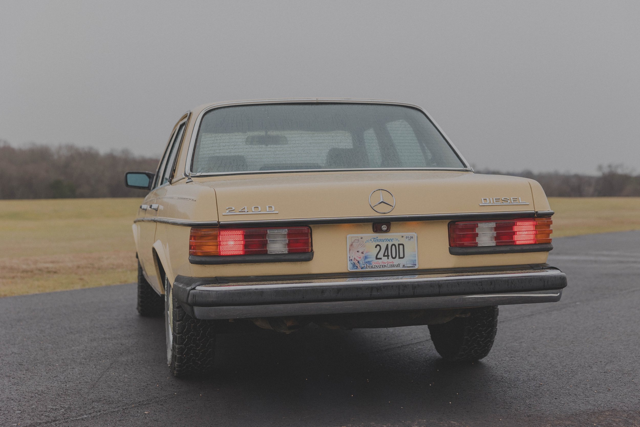 Rear view of a vintage beige Mercedes-Benz 240D with Tennessee license plate, parked on wet pavement with grassy field background.