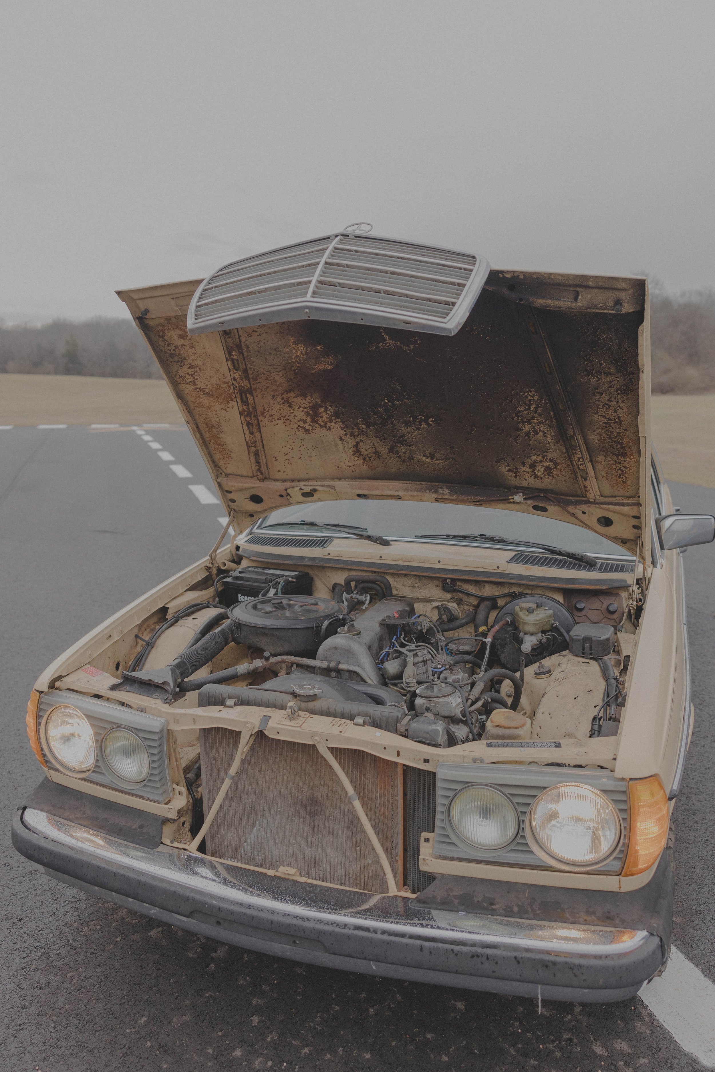 Old car with hood open showing engine and rust, parked on a road.