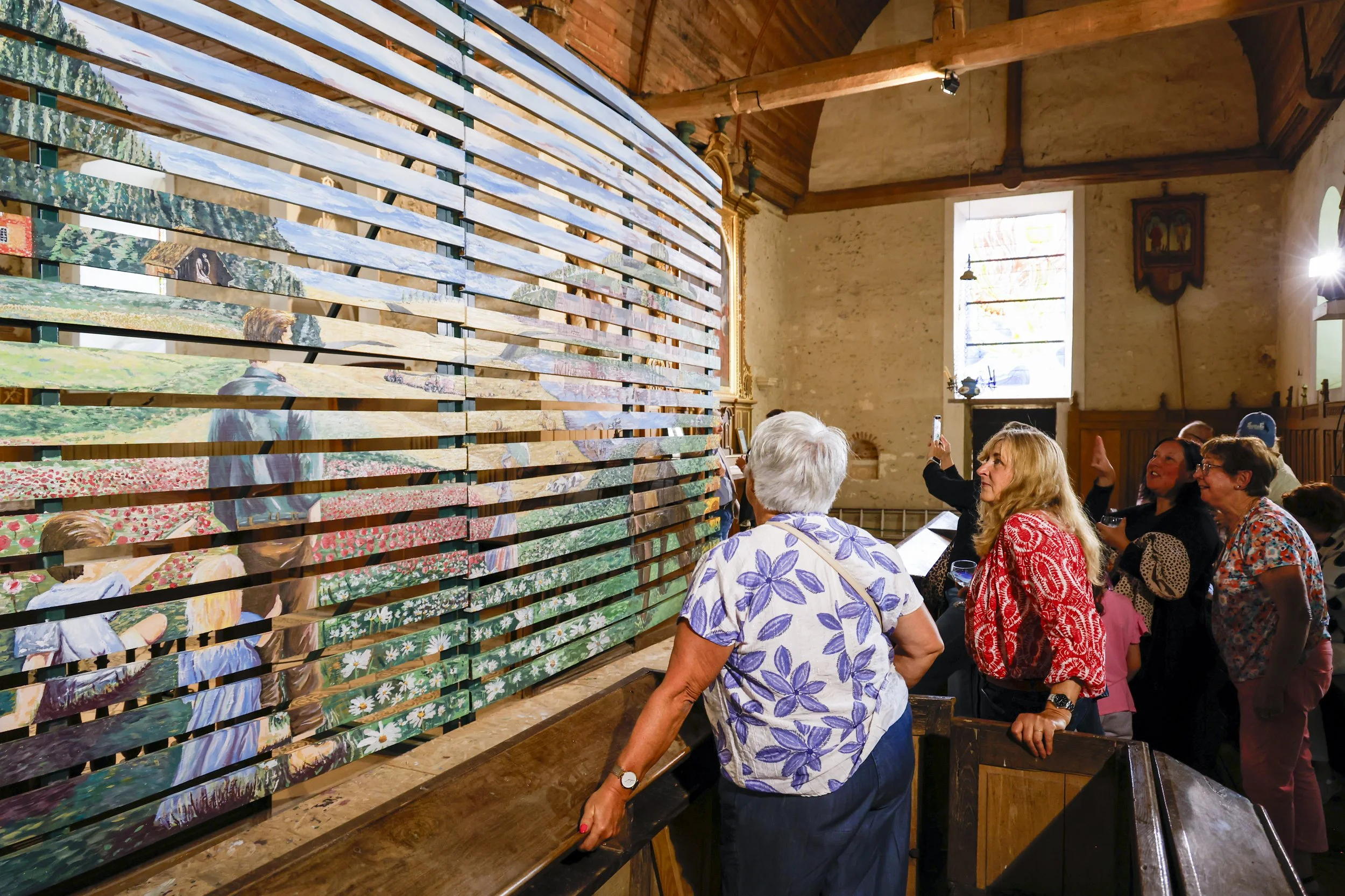 Groupe de personnes âgées admirant une œuvre d'art composée de nombreuses planches de bois peintes, exposée dans une salle avec murs en pierre et grande fenêtre laissant entrer la lumière.