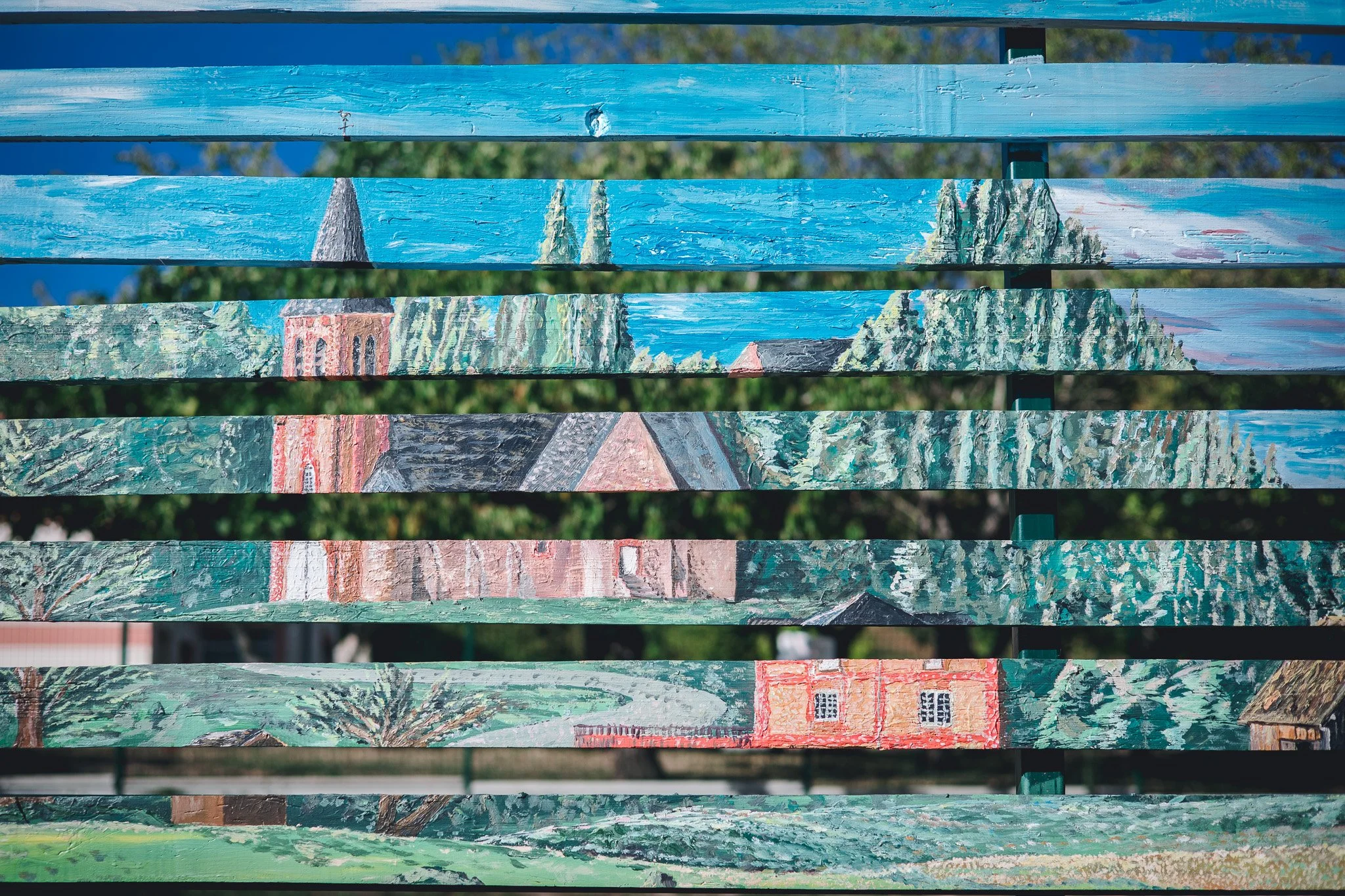 Une banderole en bois peinte avec une toile de fond de ciels bleu et de nature, représentant une scène de paysage avec des arbres, un lac, une église, et un chemin, réalisée dans un style impressionniste.