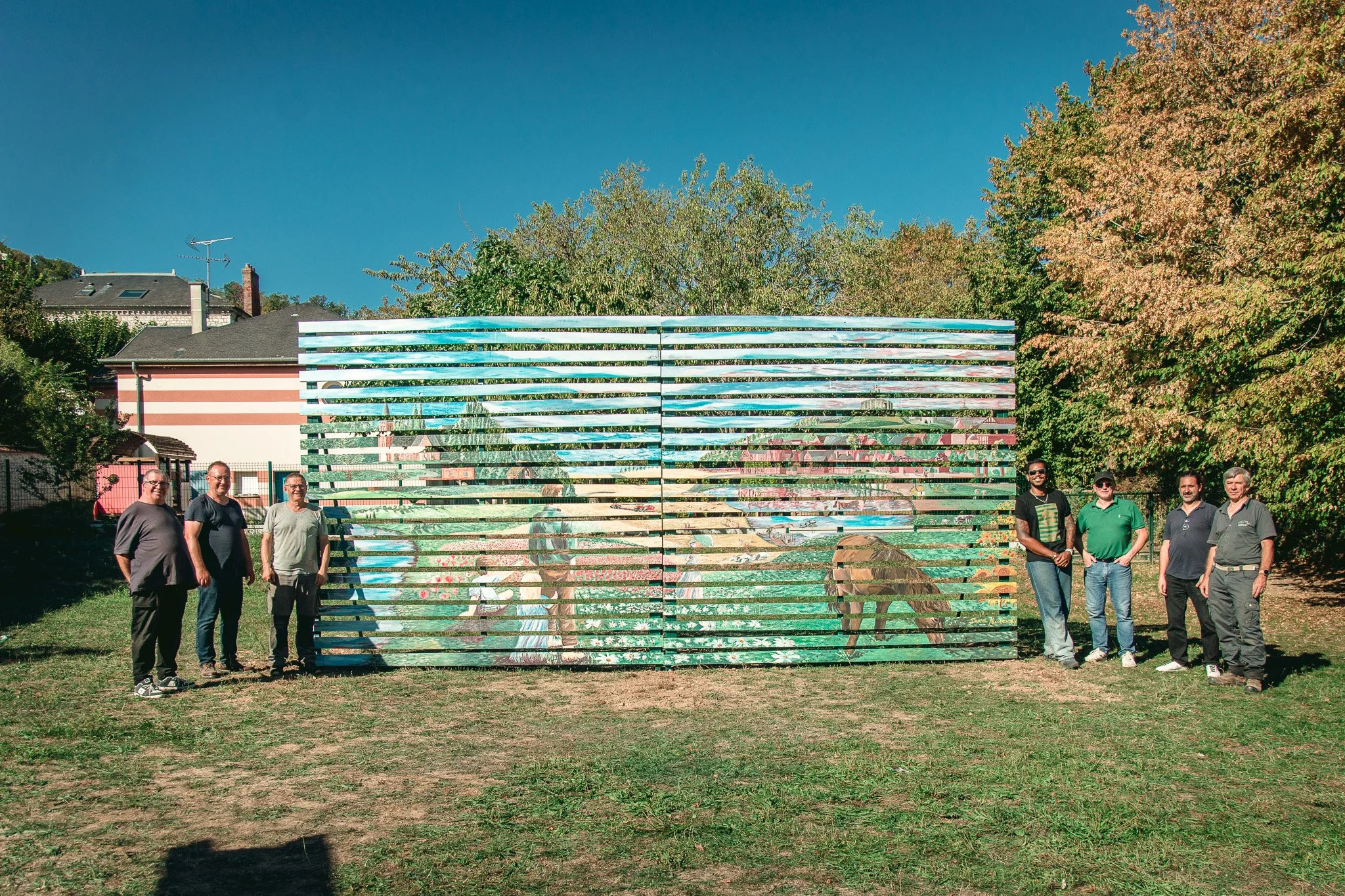 Groupe de huit personnes debout à l'extérieur devant une sculpture murale colorée en horizontal, entourée d'arbres et de maisons en arrière-plan, sous un ciel bleu.