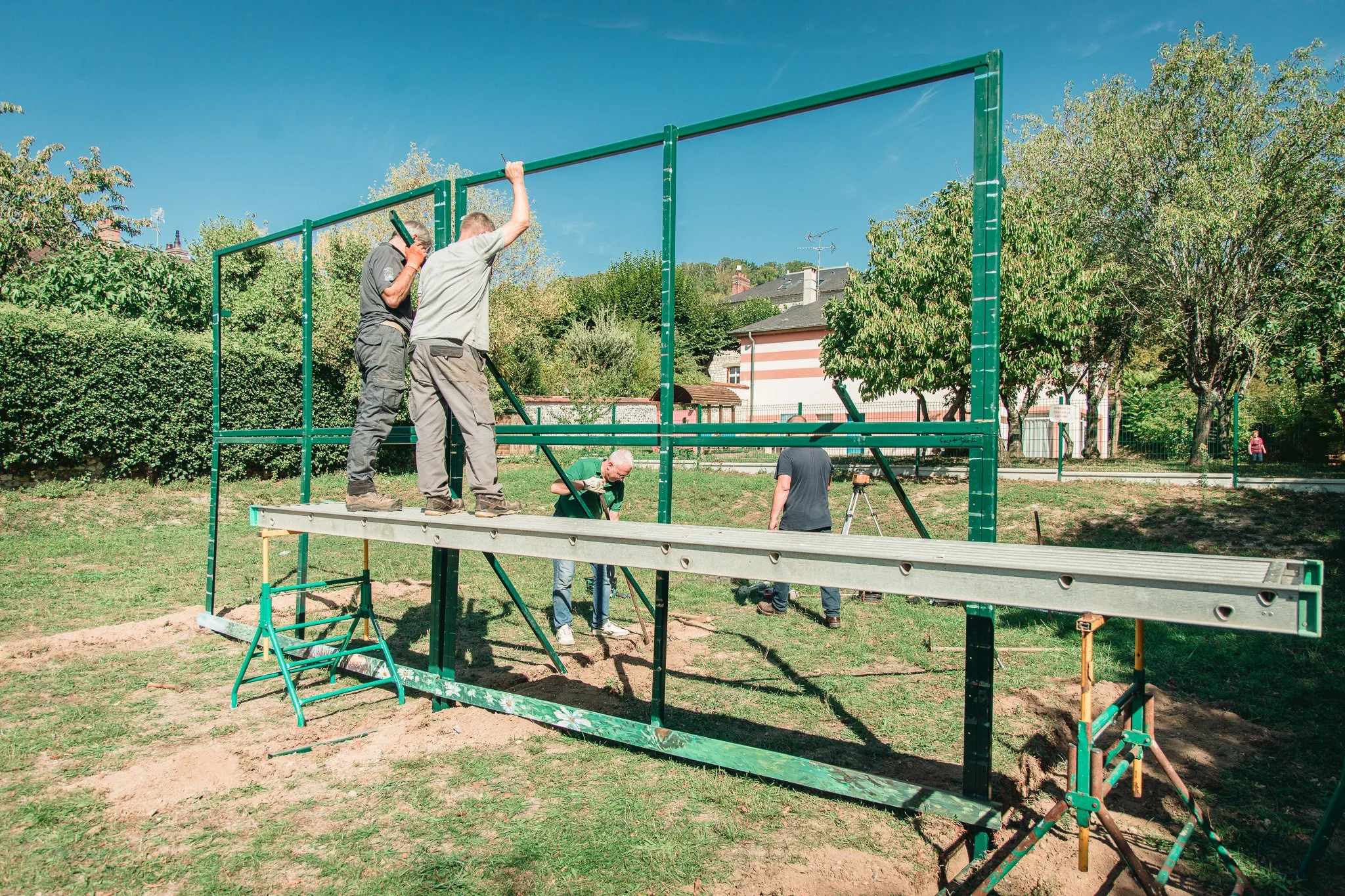 Plusieurs personnes assemblent une structure métallique en extérieur sous un ciel bleu, avec des arbres et des maisons en arrière-plan.