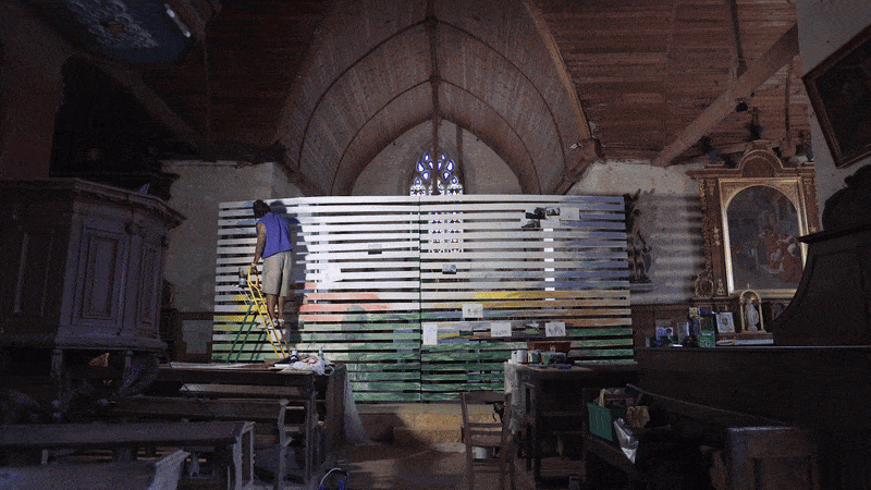 Une personne en train de peindre un mur en utilisant un escabeau dans une église avec des éléments d'ameublement ancien et une grande fenêtre en vitrail.