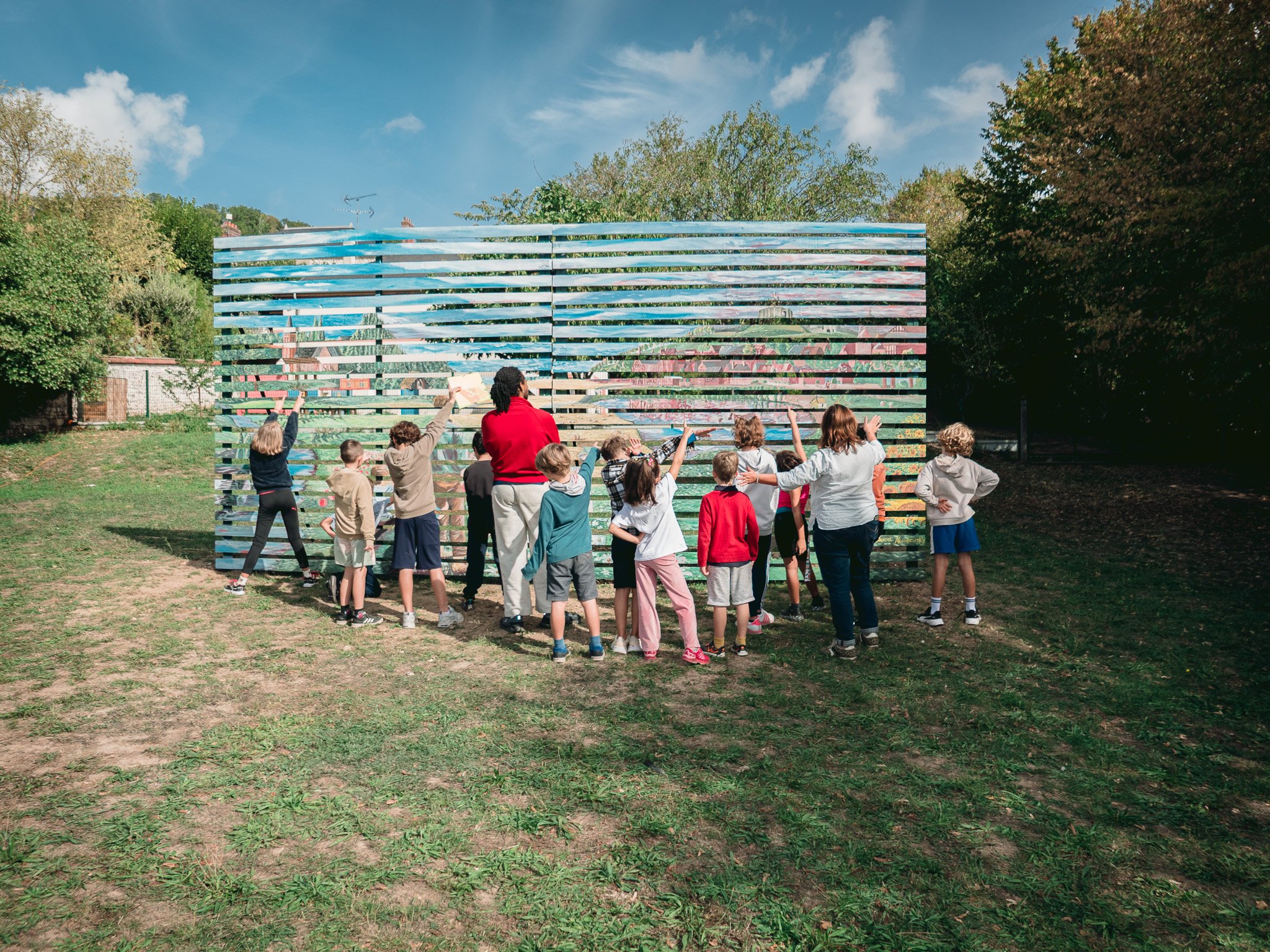 Un groupe d'enfants et un adulte se tiennent devant une clôture en bois peint avec une fresque colorée de paysages. Le ciel est bleu avec quelques nuages, et il y a des arbres autour.