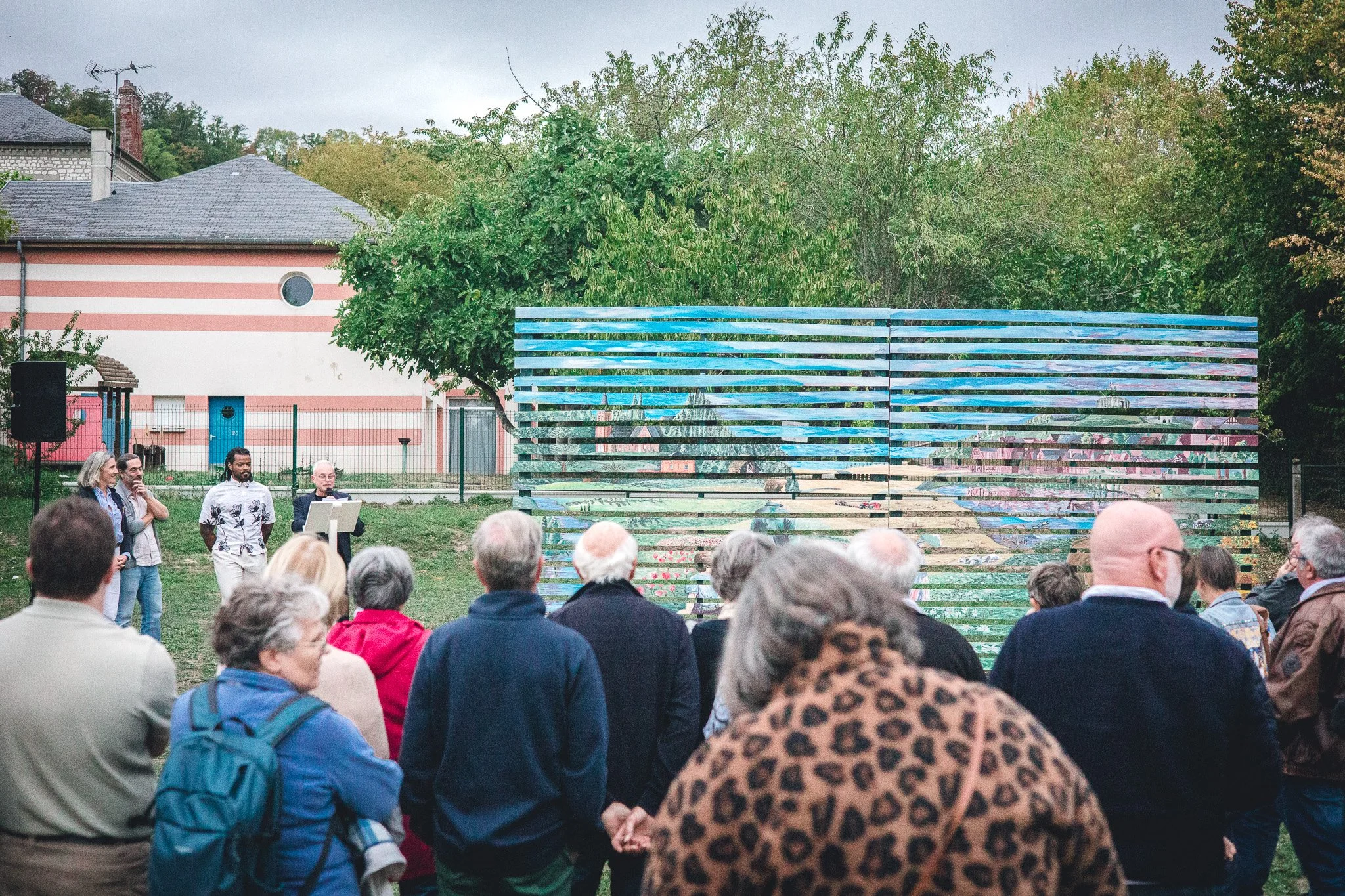 Un groupe de personnes assises et debout lors d'une réunion ou d'un événement en plein air, avec un mur coloré de graffitis en arrière-plan.