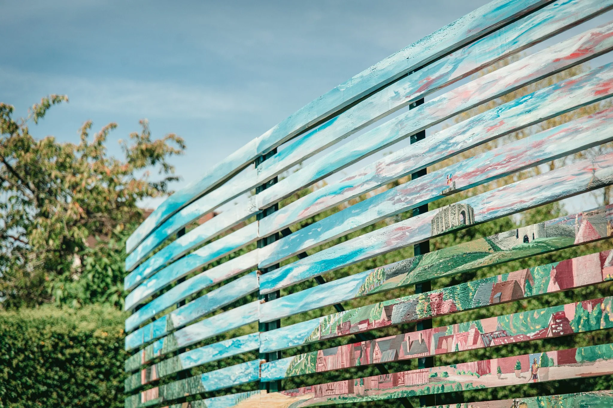 Banc en bois peint avec plusieurs couleurs, installé dans un espace extérieur avec des arbres en arrière-plan.