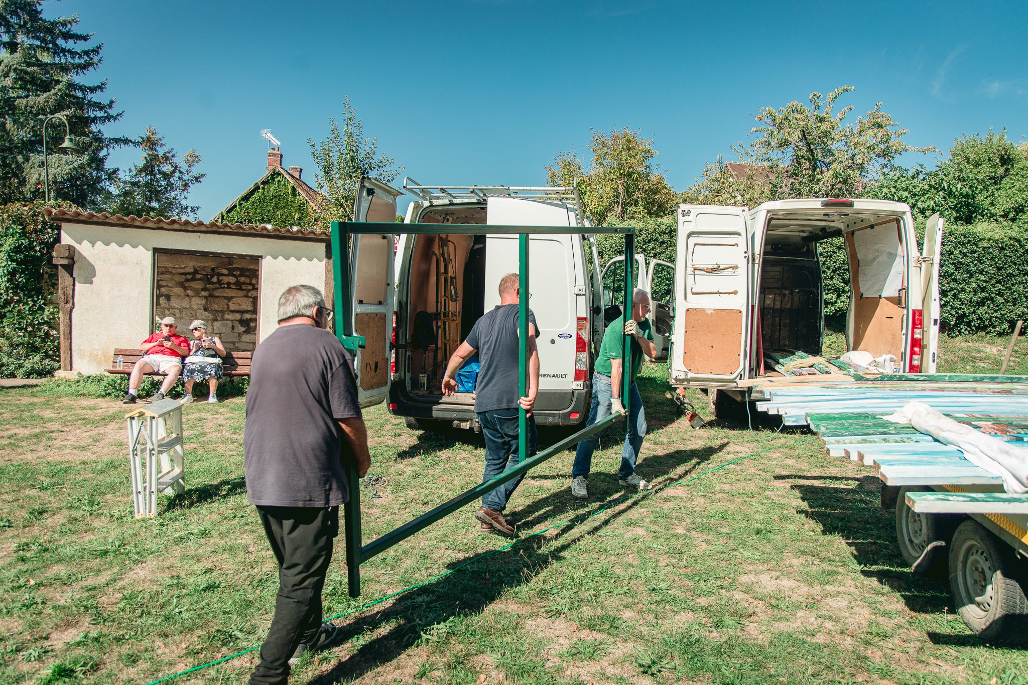 Groupe de personnes déballant et assemblant des meubles dans un jardin ensoleillé, avec deux vans ouverts.