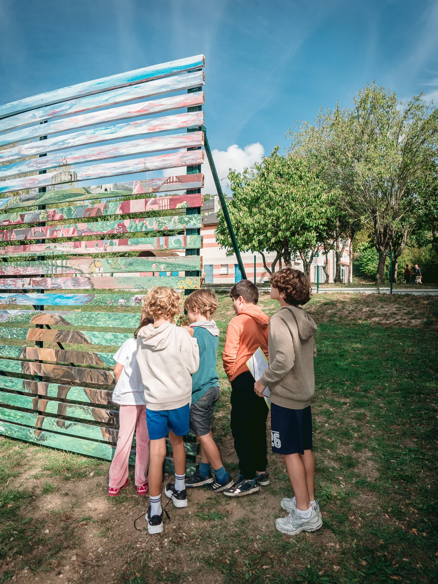 Un groupe d'enfants se tiennent devant une structure en bois peinte avec un paysage urbain et le ciel en arrière-plan. Ils semblent discuter ou observer quelque chose ensemble dans un parc.