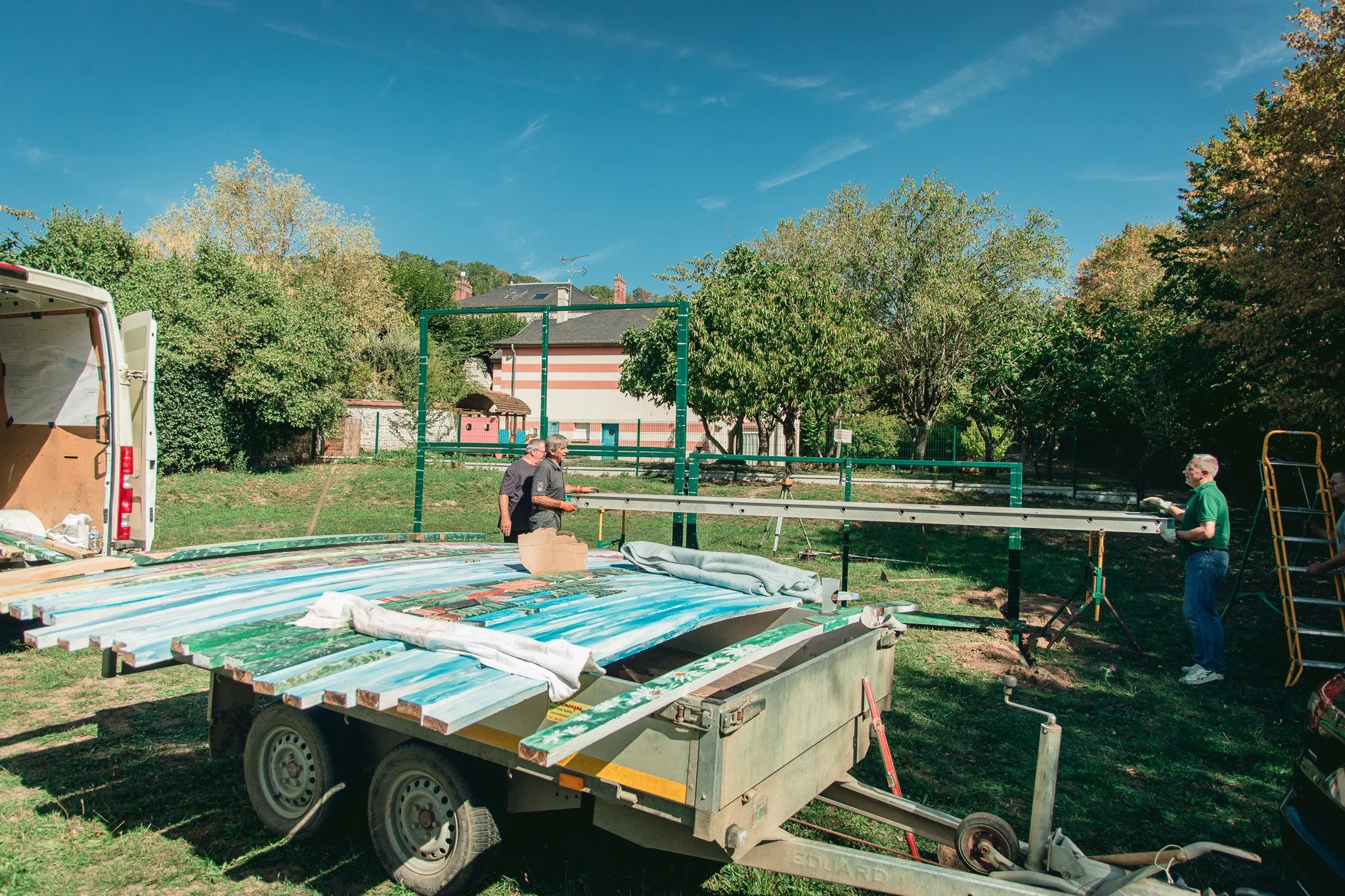 Des personnes construisent une structure en extérieu, avec des outils et des matériaux, sous un ciel ensoleillé. Sur la gauche, une camionnette ouverte avec des outils. Au centre, trois hommes concentrés sur leur tâche. En arrière-plan, des arbres et
