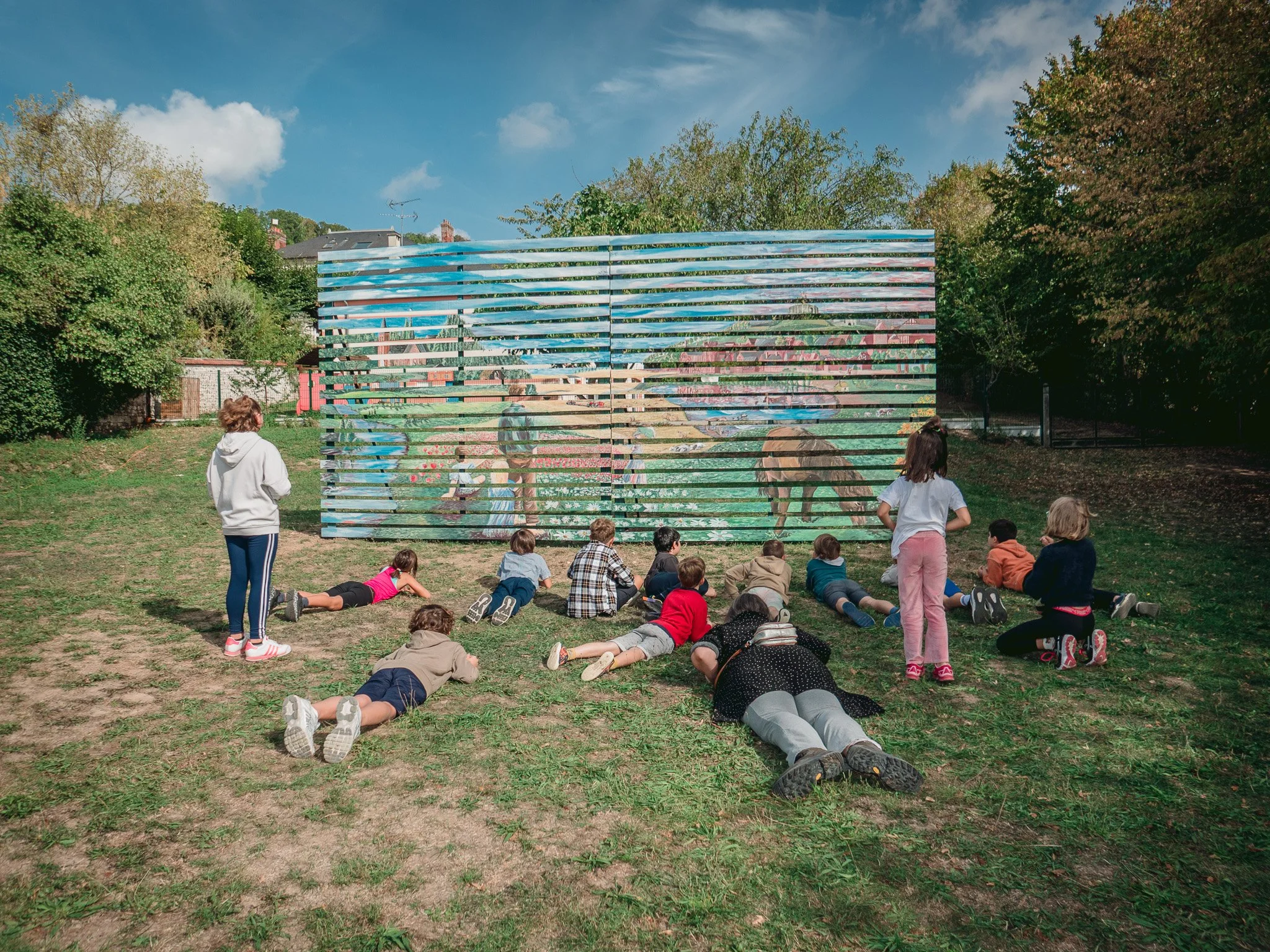 Un groupe d'enfants et d'adultes dans un parc, certains allongés sur l'herbe, observant une grande œuvre d'art mural composée de bandes horizontales reflectives, avec des arbres et un ciel bleu en arrière-plan.