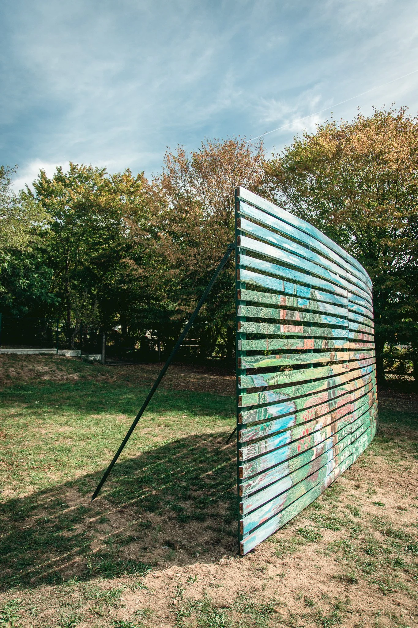 Une sculpture en forme de mur incurvé composée de planches peintes de couleurs variées, située dans un parc avec des arbres en arrière-plan et un ciel bleu avec quelques nuages.