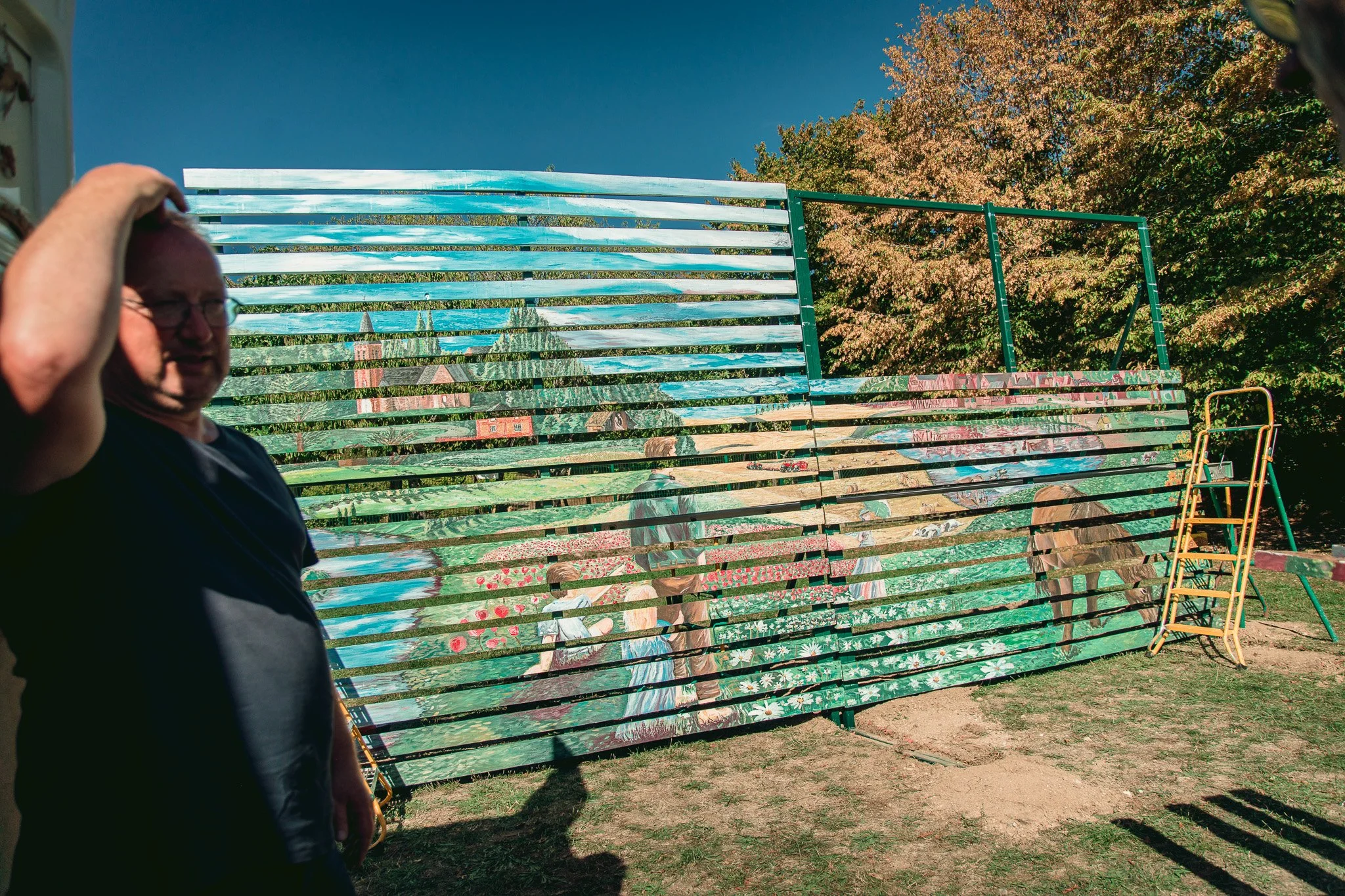 Une personne moustachue portant des lunettes, en t-shirt noir, à gauche de l'image, avec un fond comprenant une grande structure métallique peinte avec une peinture murale représentant un paysage avec des maisons, un ciel bleu, des arbres en été, et 