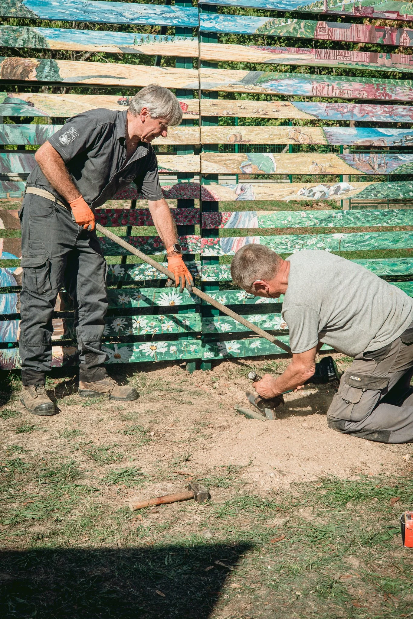 Deux hommes travaillent au sol avec des outils à l'extérieur, devant un mur en bois peint avec des scènes agricoles et naturelles.