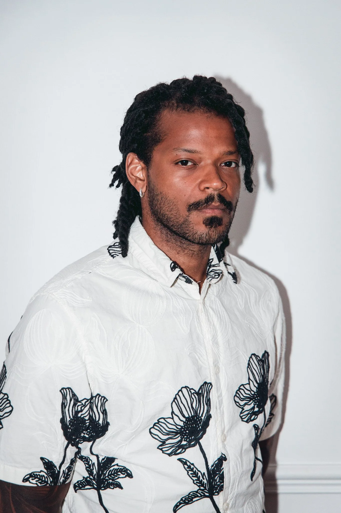 Portrait d'un homme avec des cheveux bouclés en dreadlocks, portant une chemise blanche avec des motifs de fleurs noires.